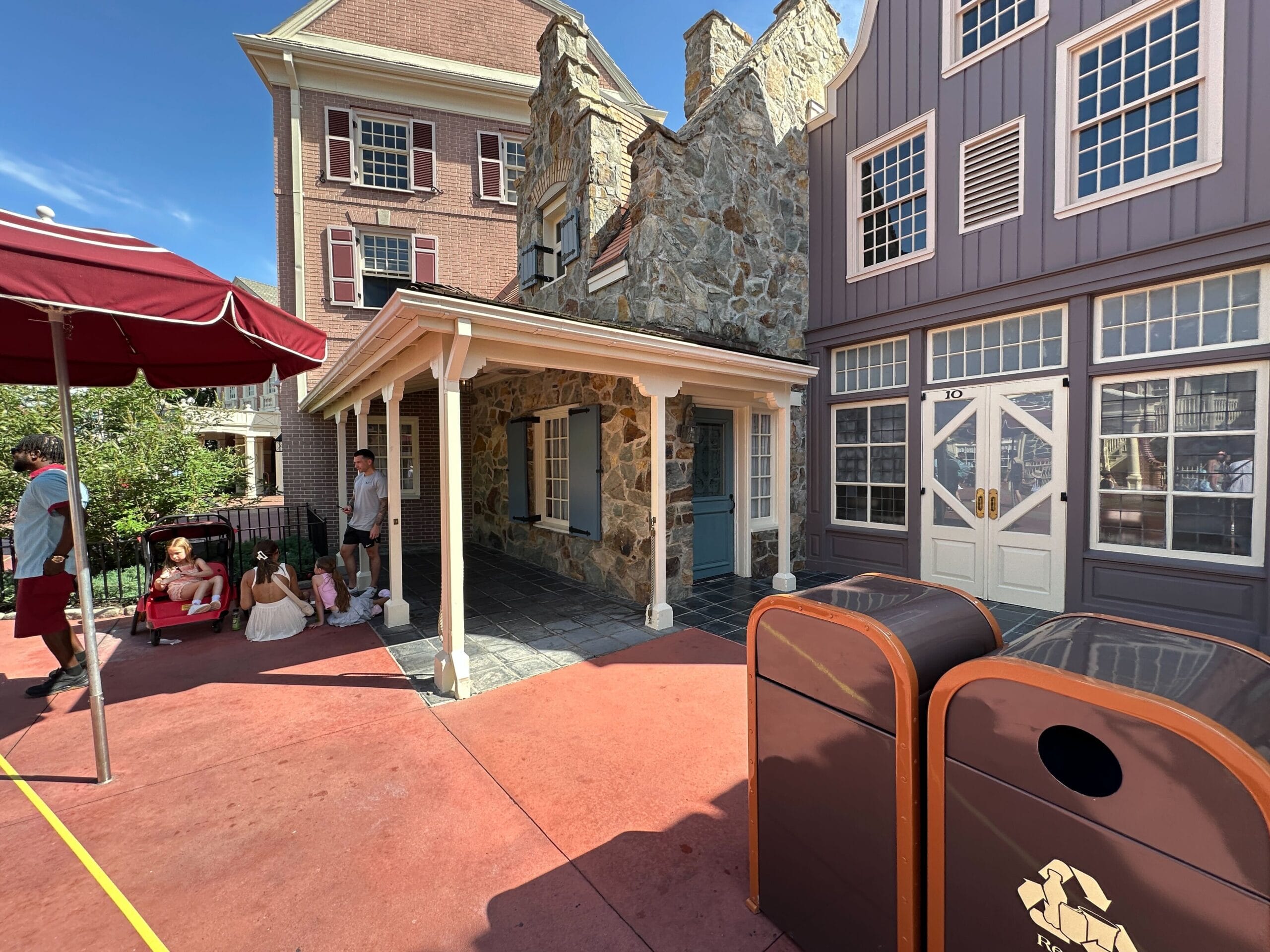 People rest in the shade beside stone and wooden colonial-style buildings with two brown trash bins in the foreground.