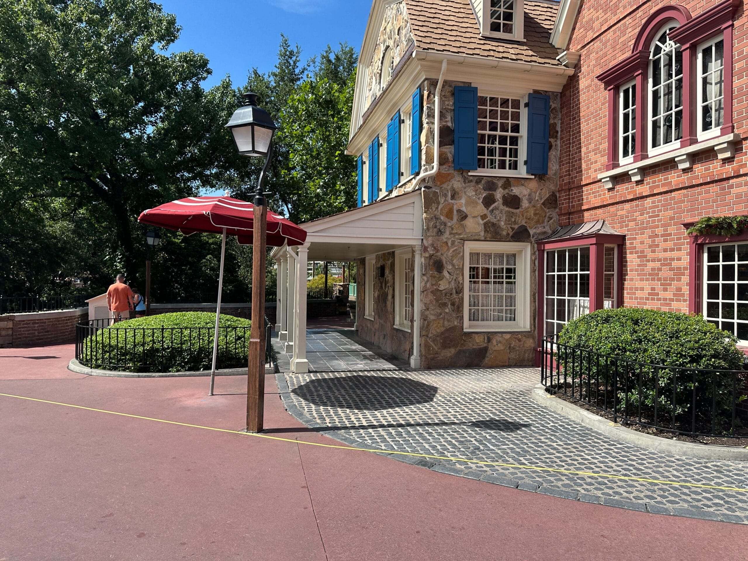 A sunny walkway passes a stone and brick building with blue shutters and a red umbrella near a streetlamp.
