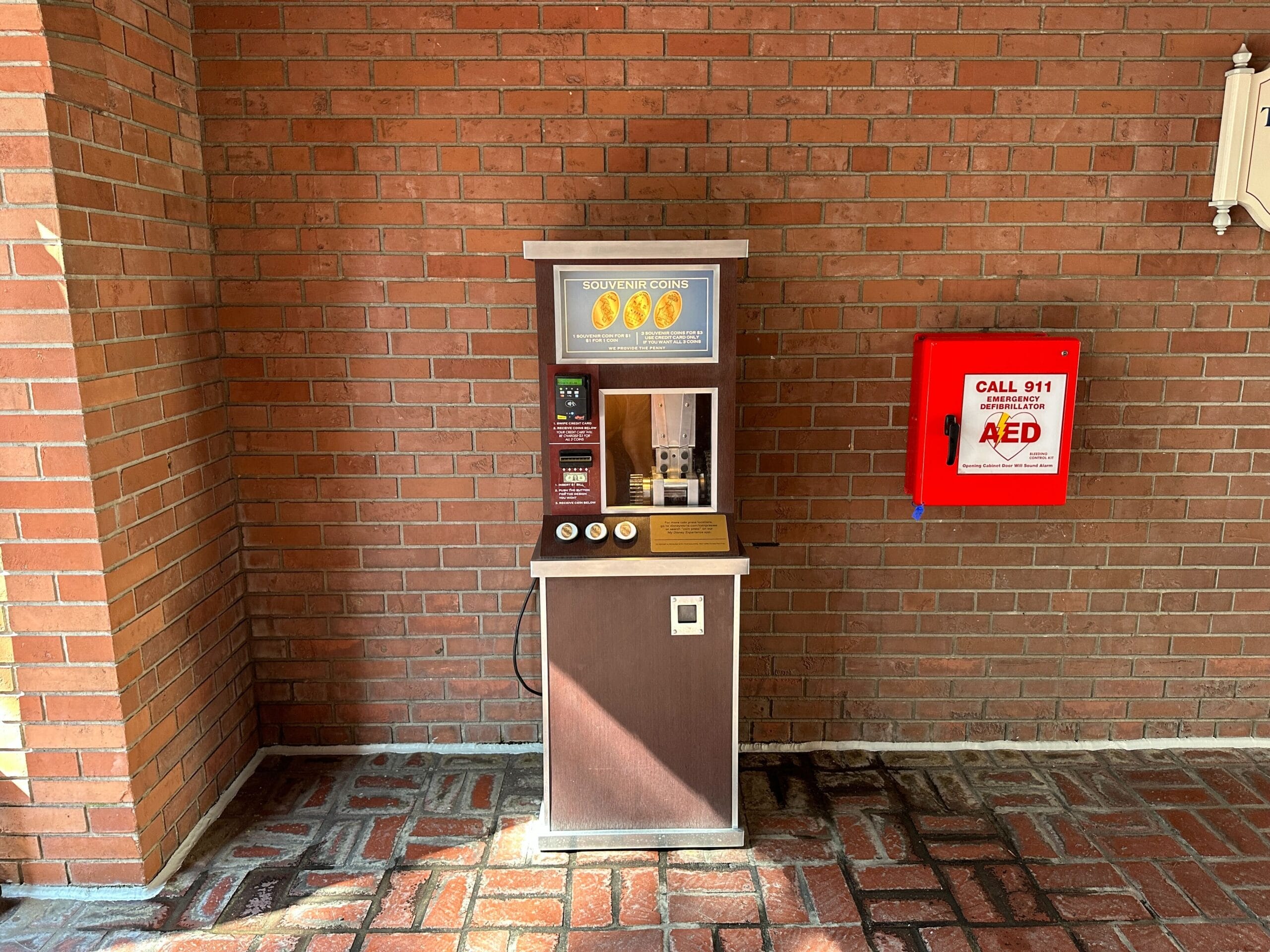 A coin press machine and a red AED box are mounted on a brick wall in a covered area.