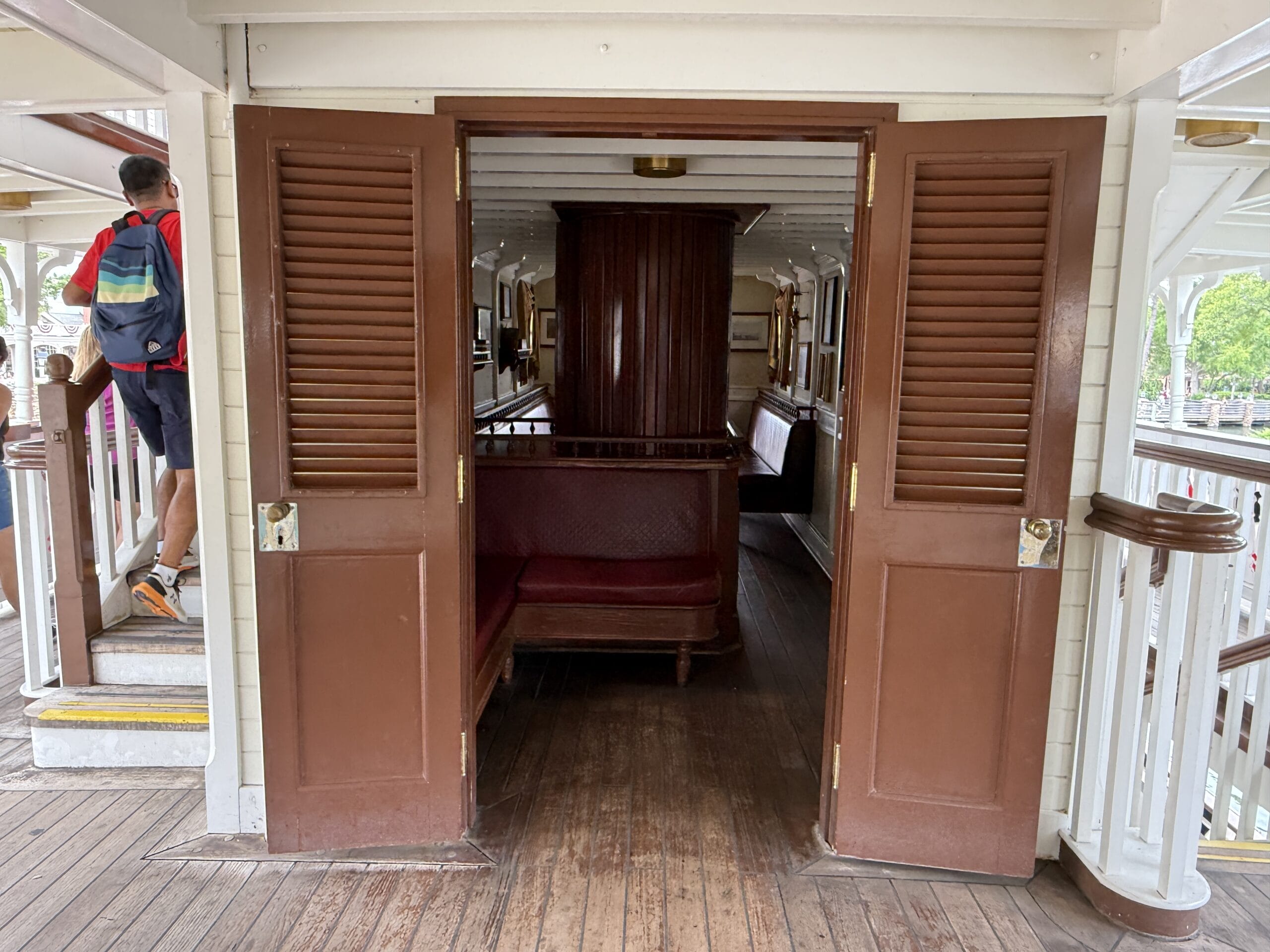 Open brown double doors reveal a wooden bench; a person with a backpack walks upstairs like boarding the Liberty Square Riverboat.