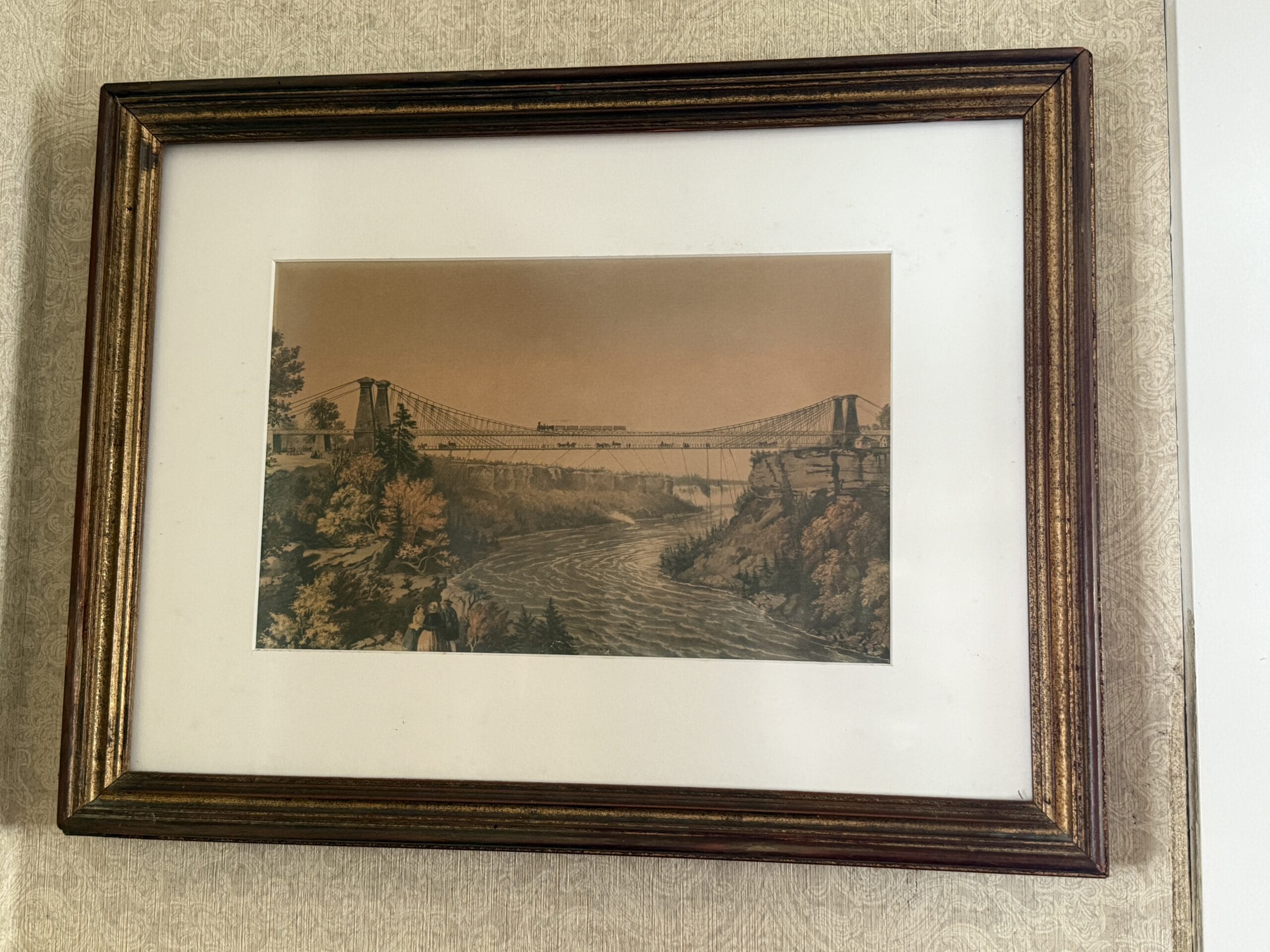 Framed painting of a suspension bridge over a river, evoking the charm of Liberty Square Riverboat, on a patterned wall.