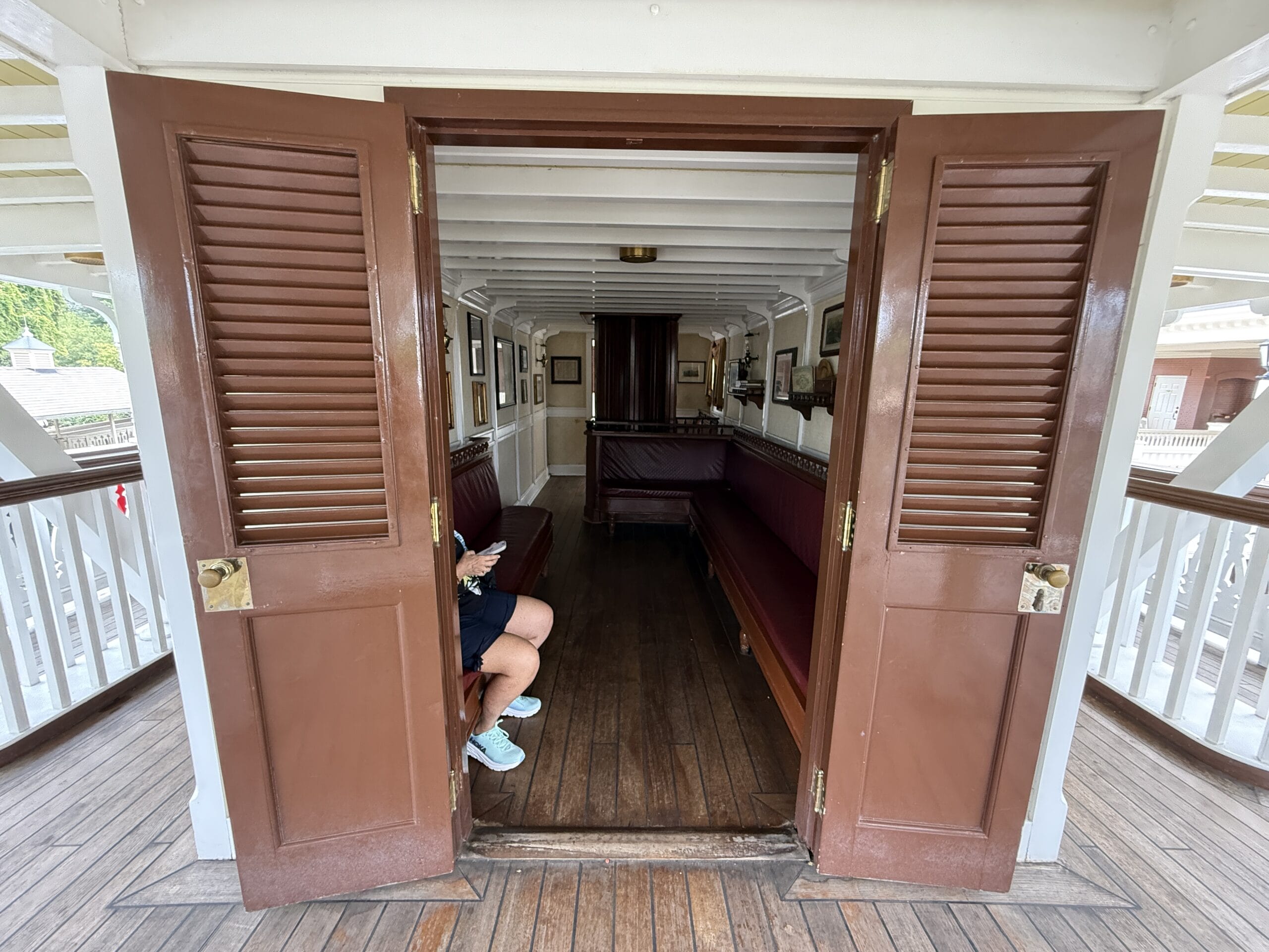 Brown double doors open to a vintage Liberty Square Riverboat cabin; a person sits inside, only legs visible, holding a phone.