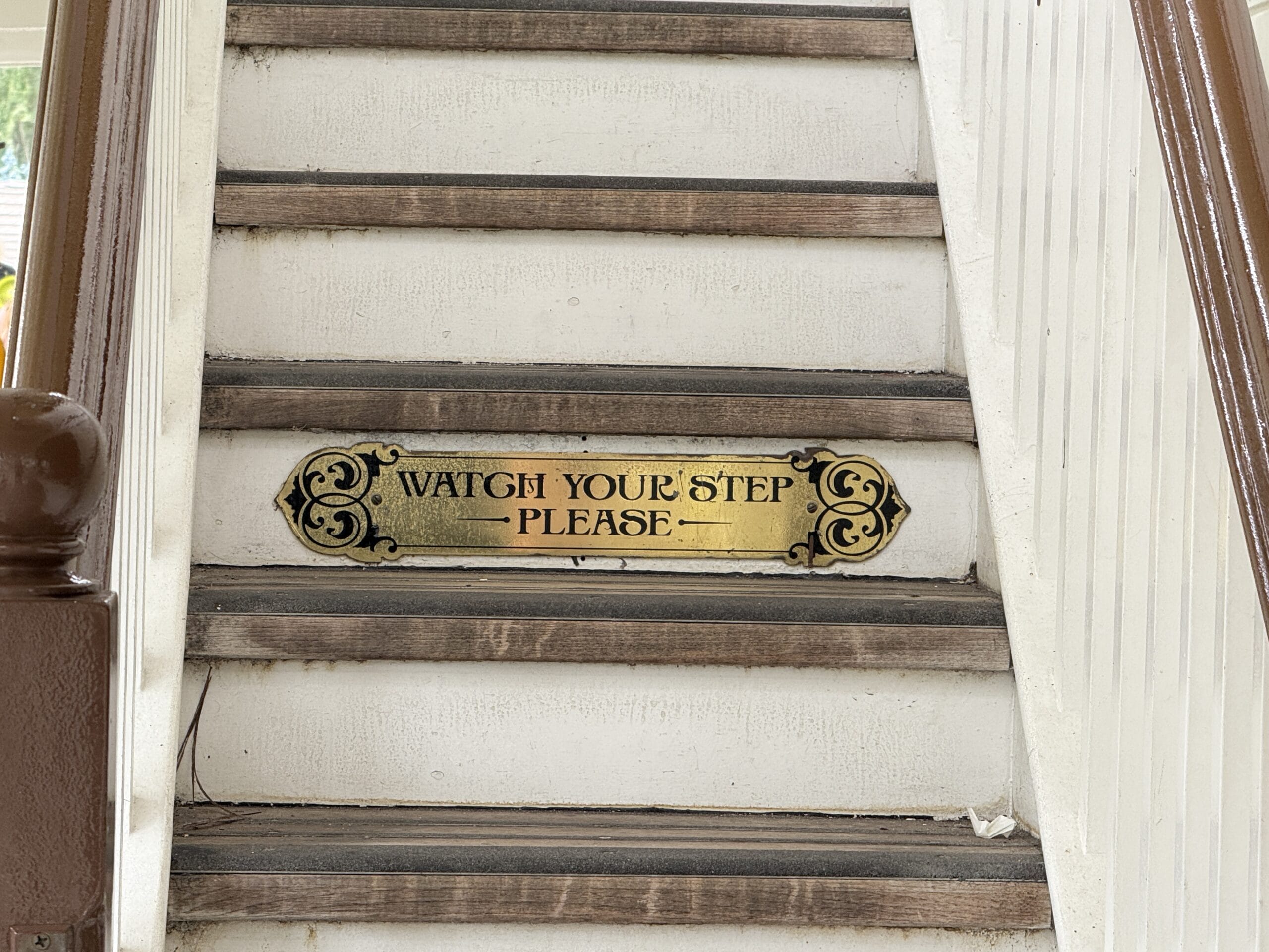 A sign on the Liberty Square Riverboat staircase at Magic Kingdom reads "Watch Your Step Please" in decorative lettering.
