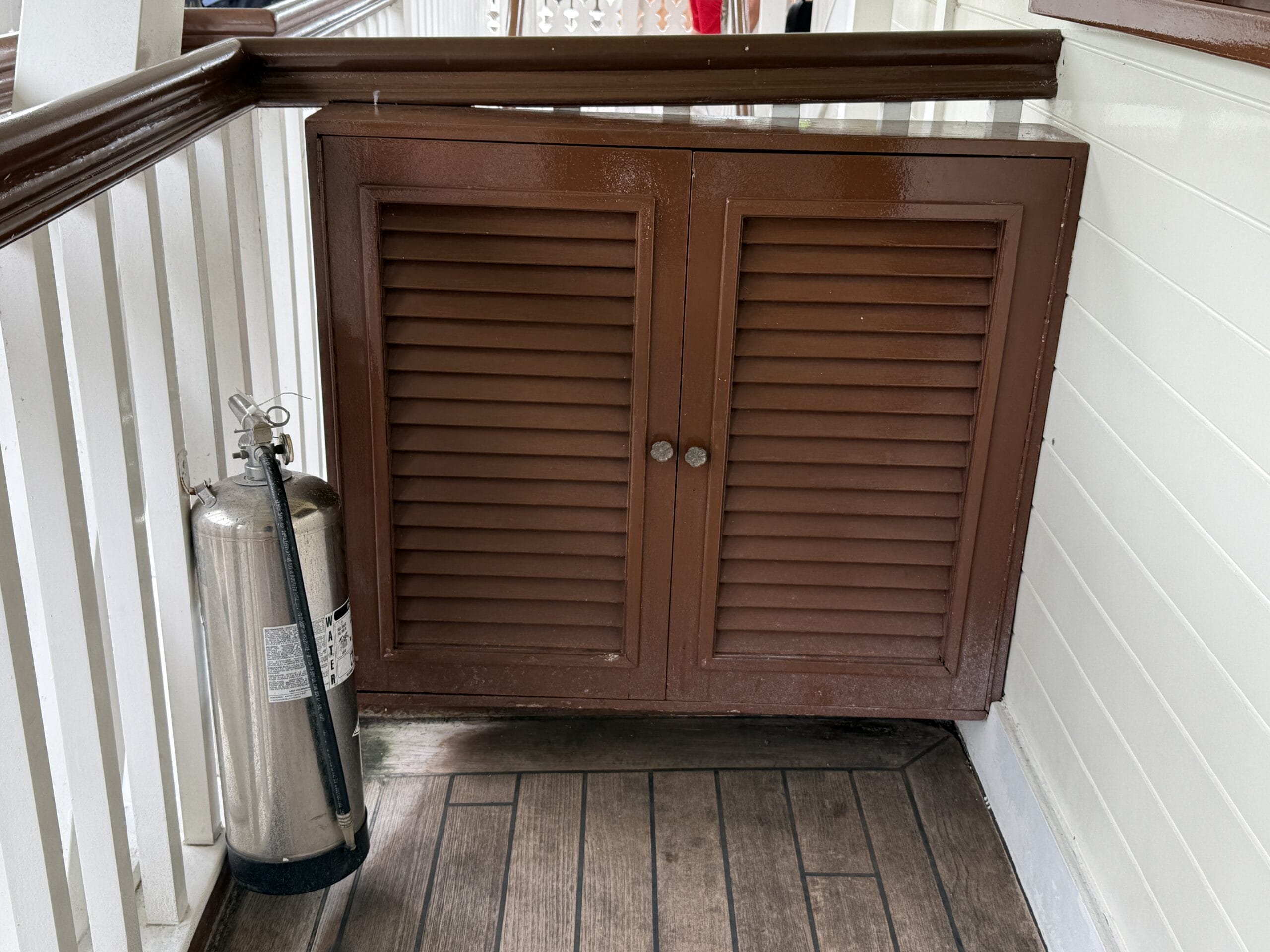 A silver fire extinguisher stands near a brown cabinet on a wooden deck by the Liberty Square Riverboat railing.