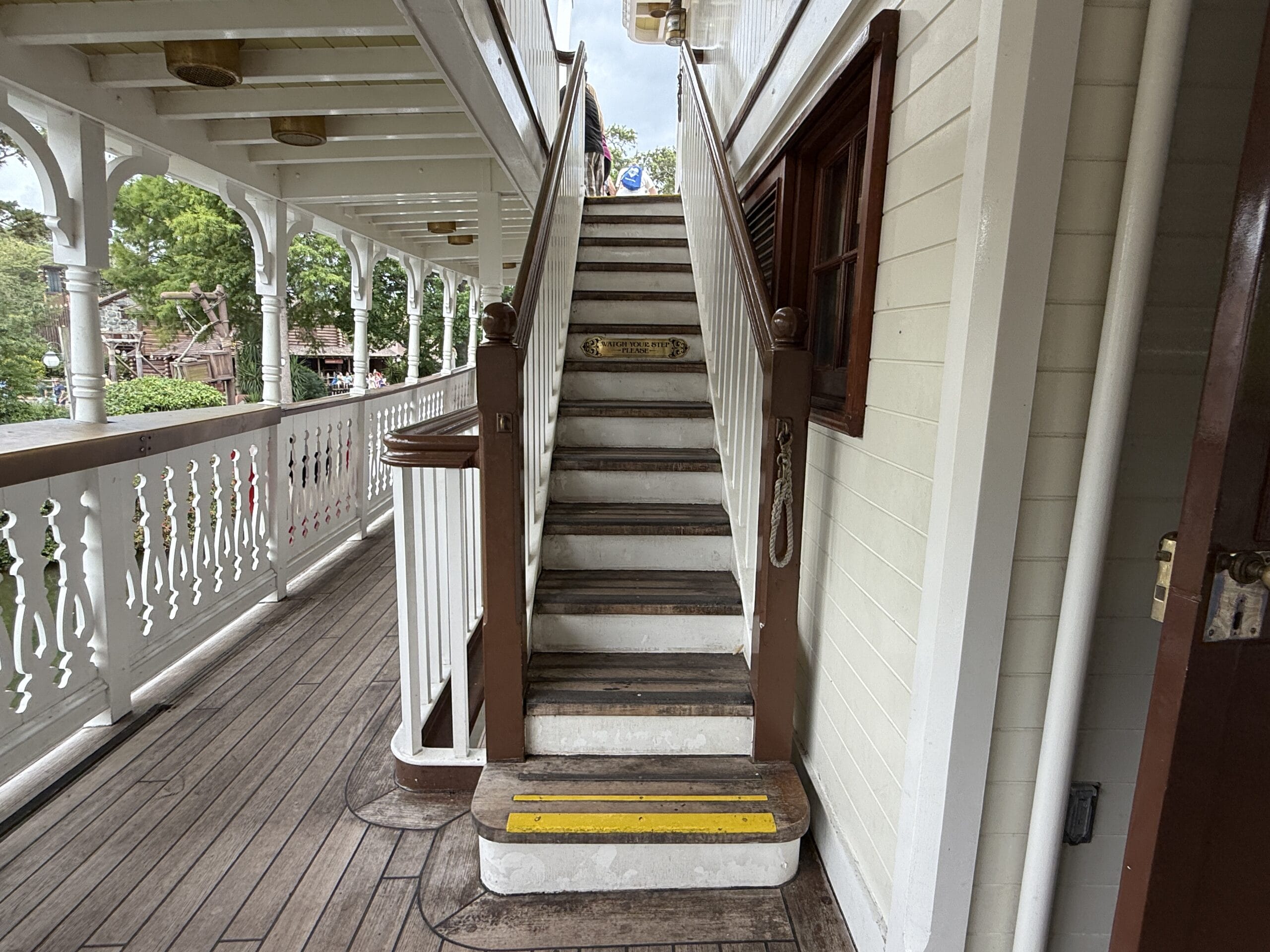 Wooden staircase with brown railings on the Liberty Square Riverboat, featuring a yellow caution strip at the base.