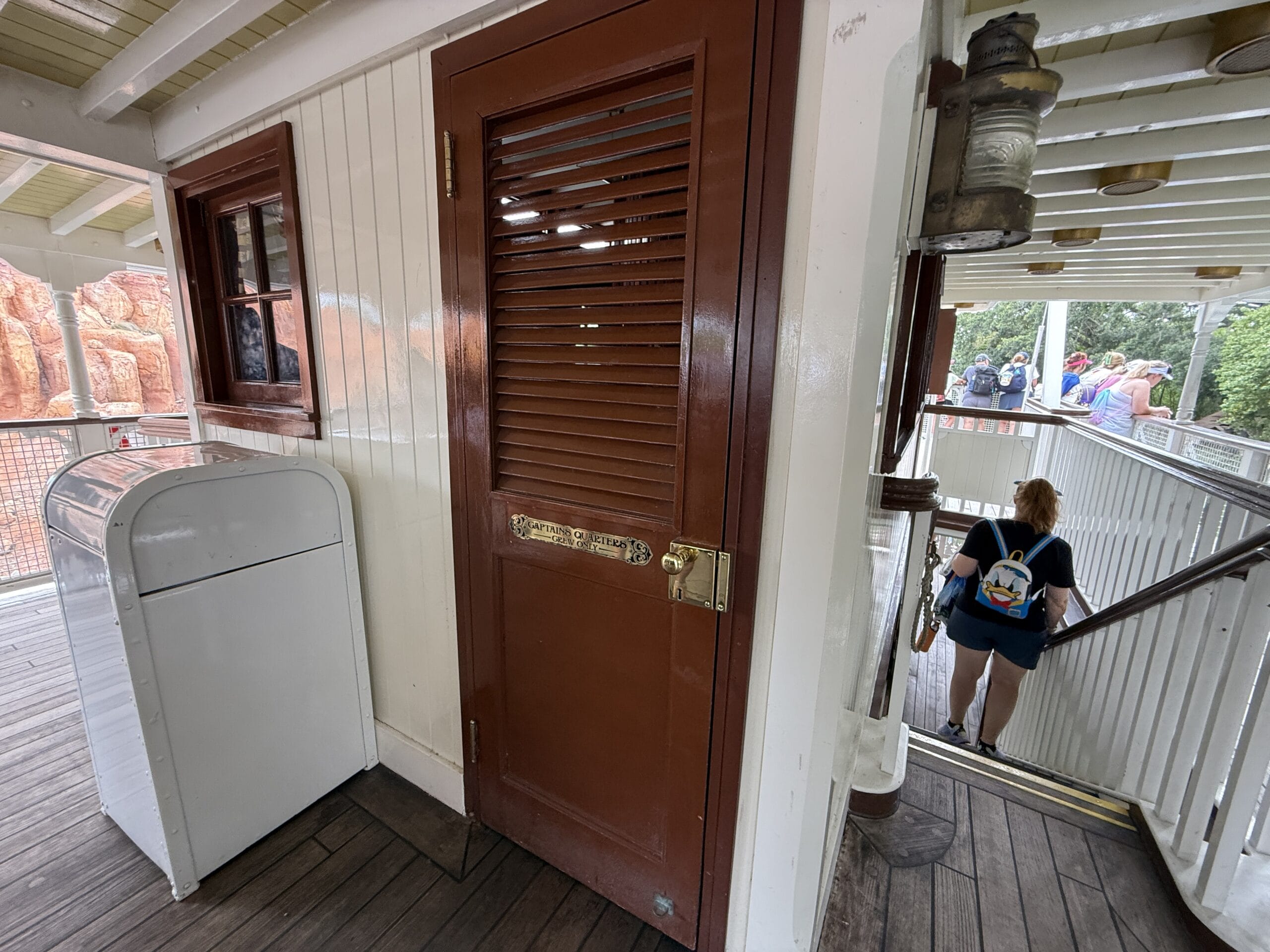 A brown louvered door on a wooden deck near a white trash bin by the Liberty Square Riverboat, with people on stairs nearby.