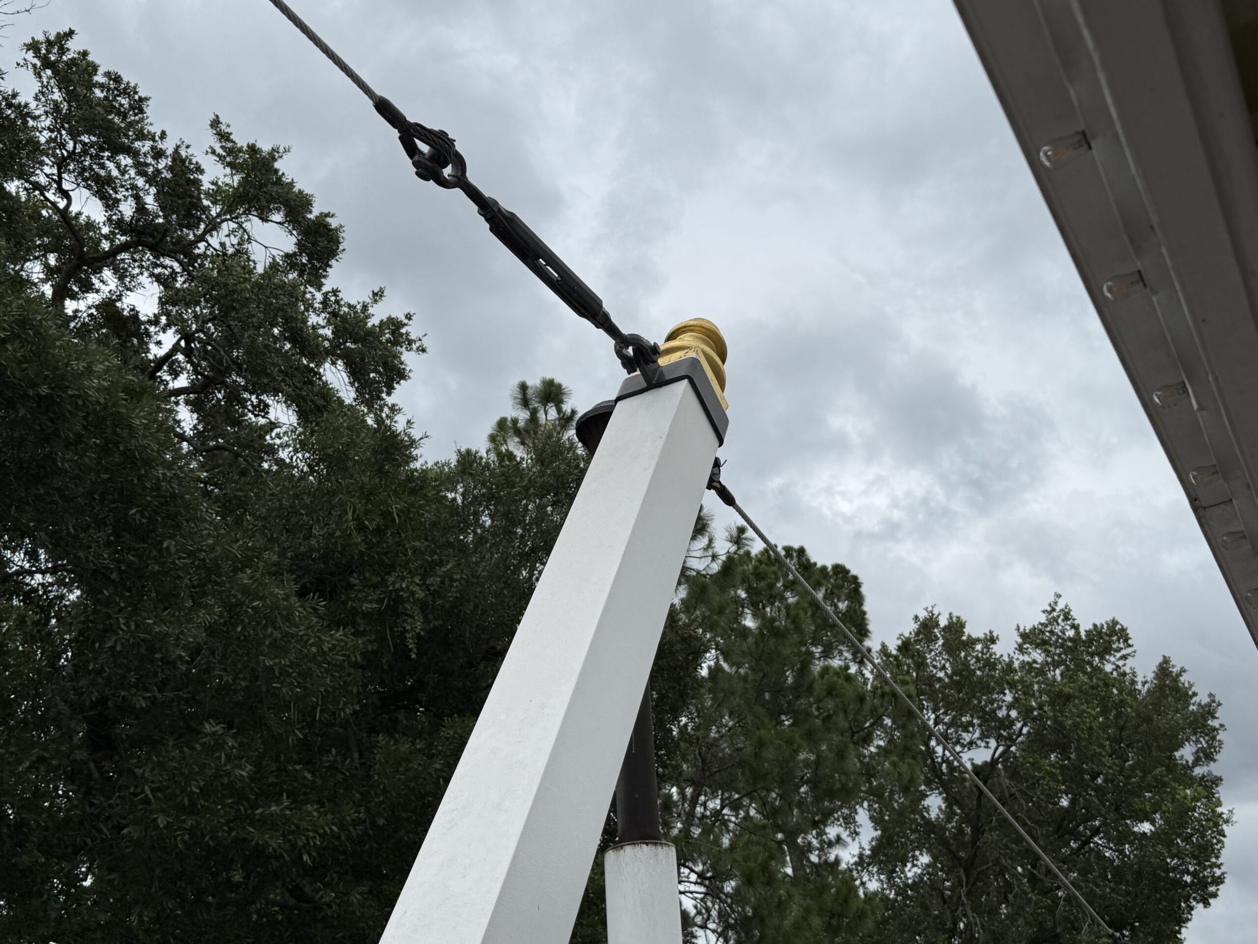 White post with guy wire and gold cap near trees, cloudy sky, and roof edge—seen by the Liberty Square Riverboat.