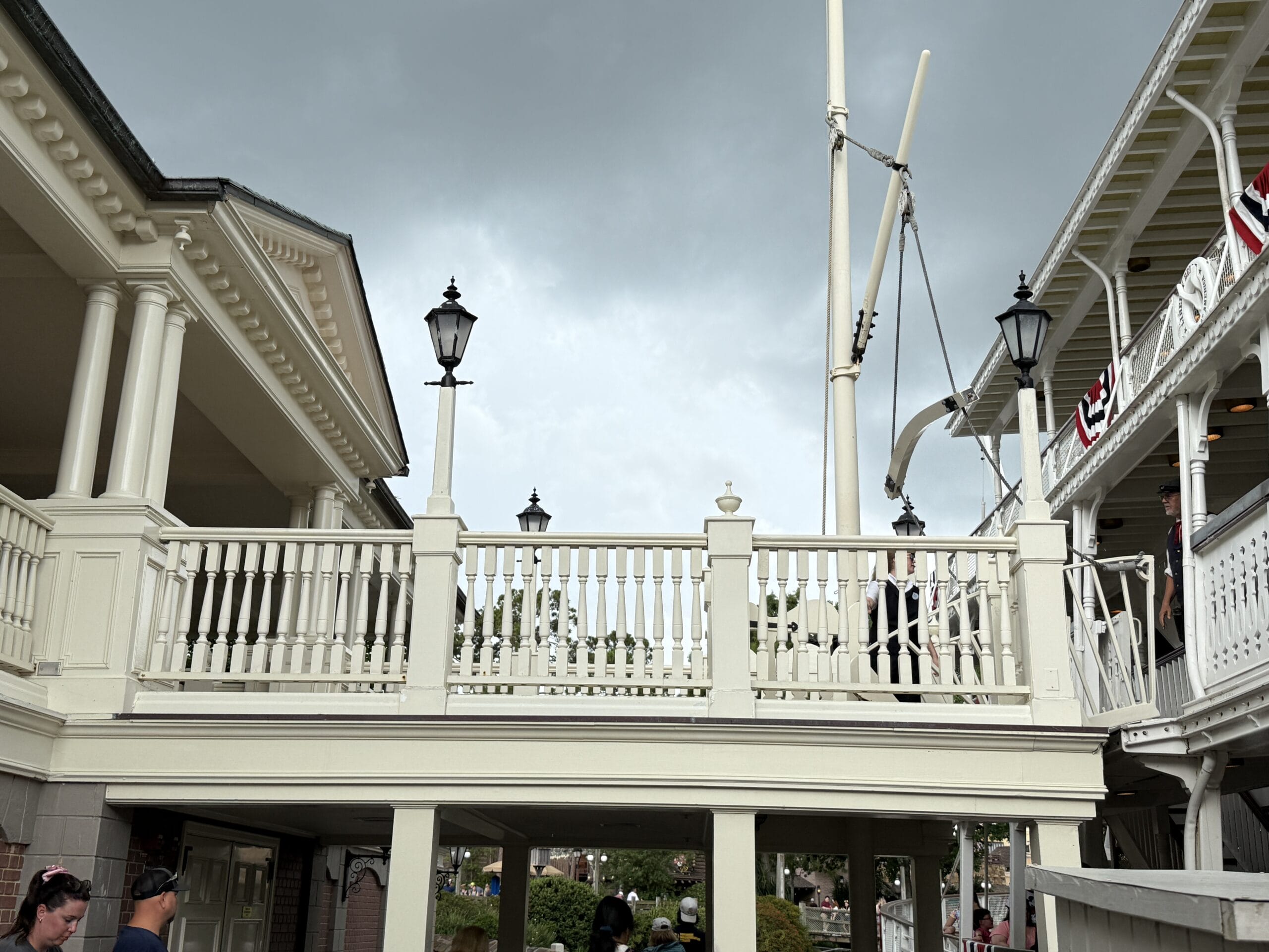 A white wooden bridge links two buildings under cloudy skies, as people stroll below and the Liberty Square Riverboat sails by.