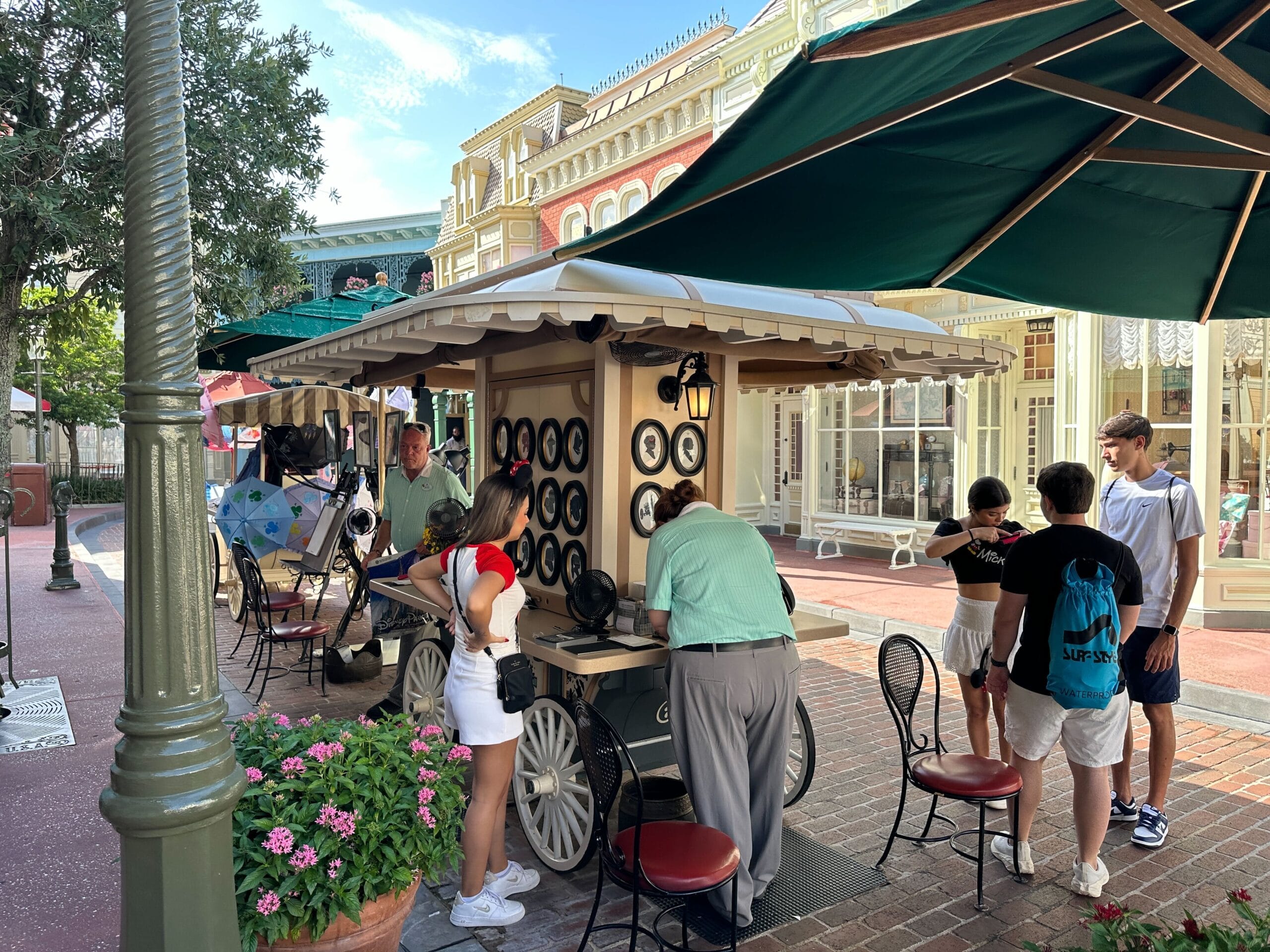 People gather around an outdoor caricature artist’s stand on a brick street with shops and green umbrellas nearby.