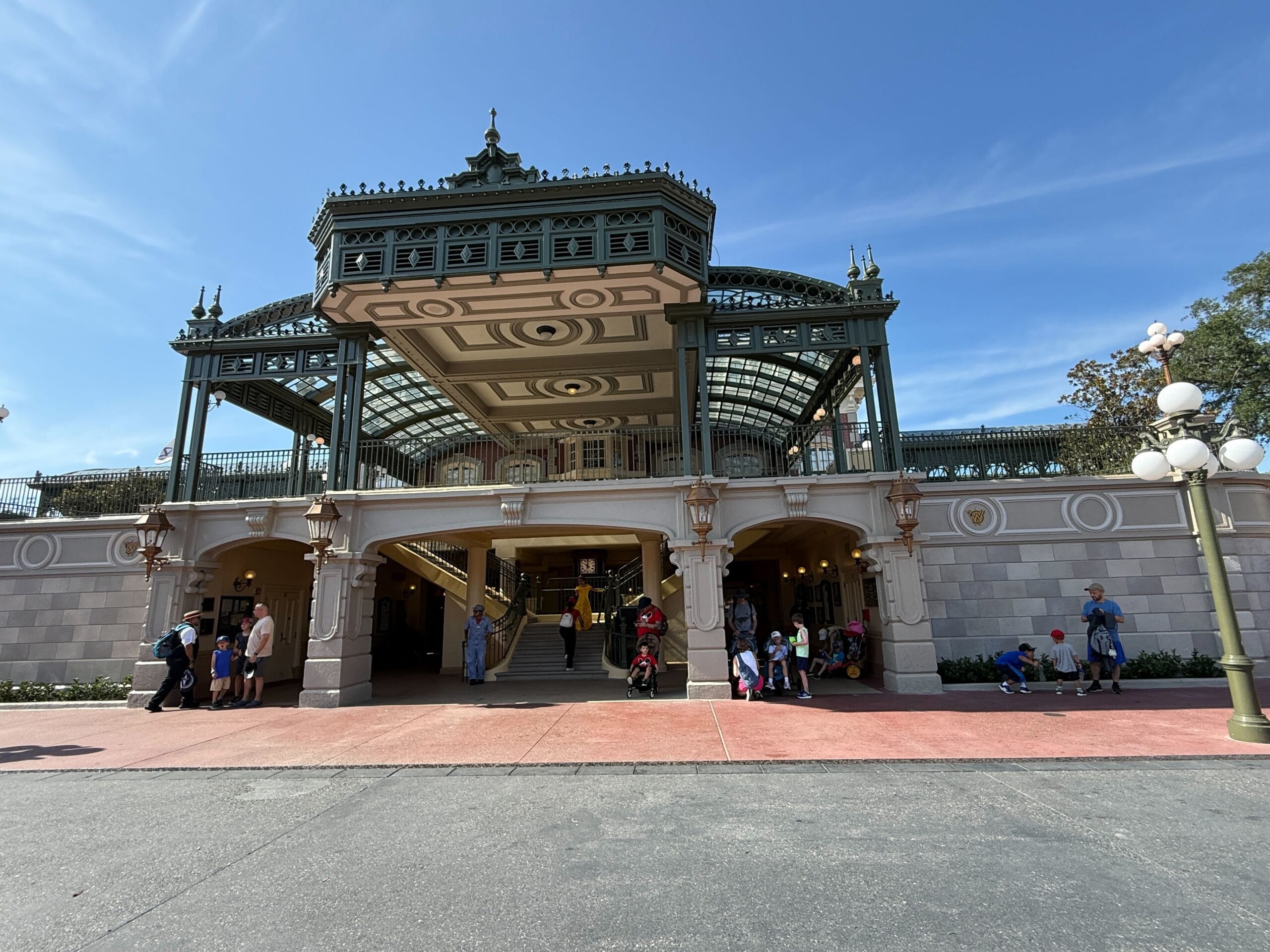 People stand and sit in front of a large, ornate building with green trim and a glass roof under a blue sky.