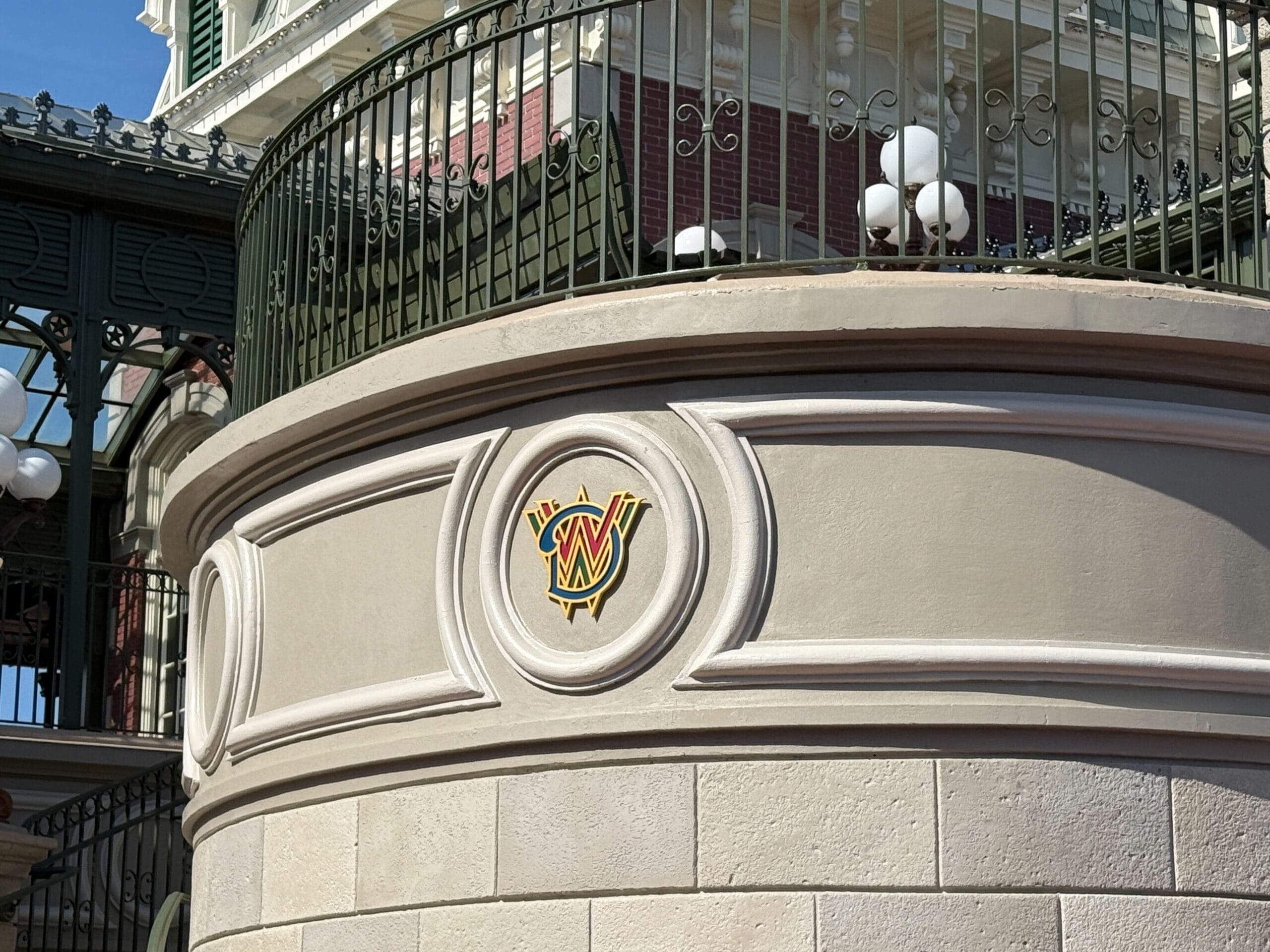 Round decorative emblem with the letters "MW" on a beige wall, next to a green railing and ornate building.