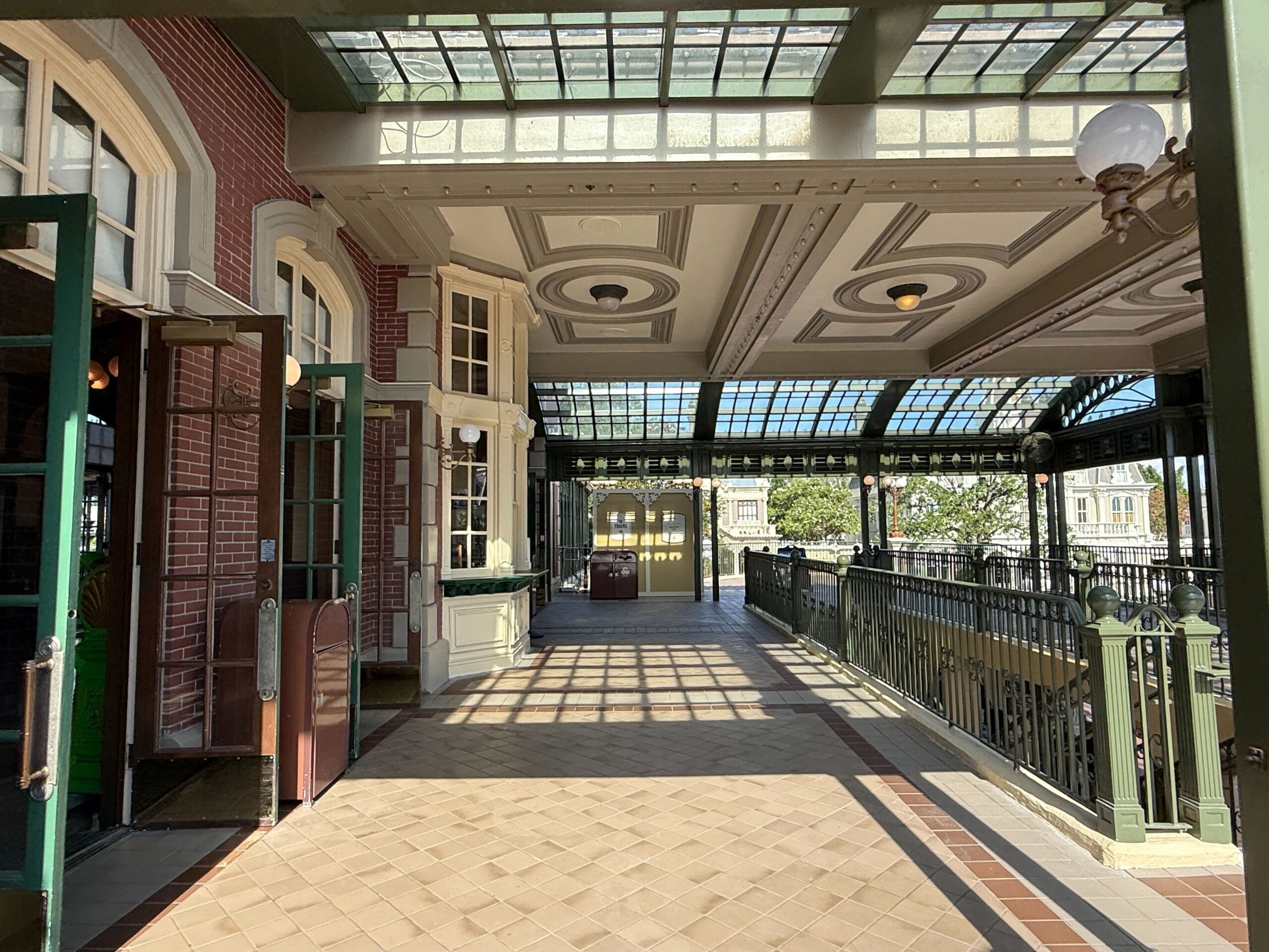 Covered outdoor walkway with glass ceiling, decorative railings, and sunlight casting shadows on tiled floor.