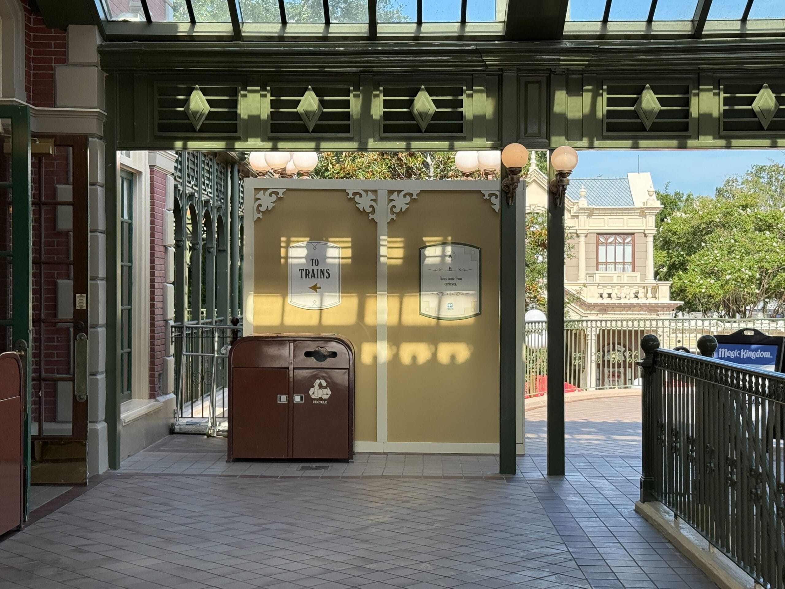 Sunlit train station platform with a brown trash can, signs, and railing, with trees and a building in the background.