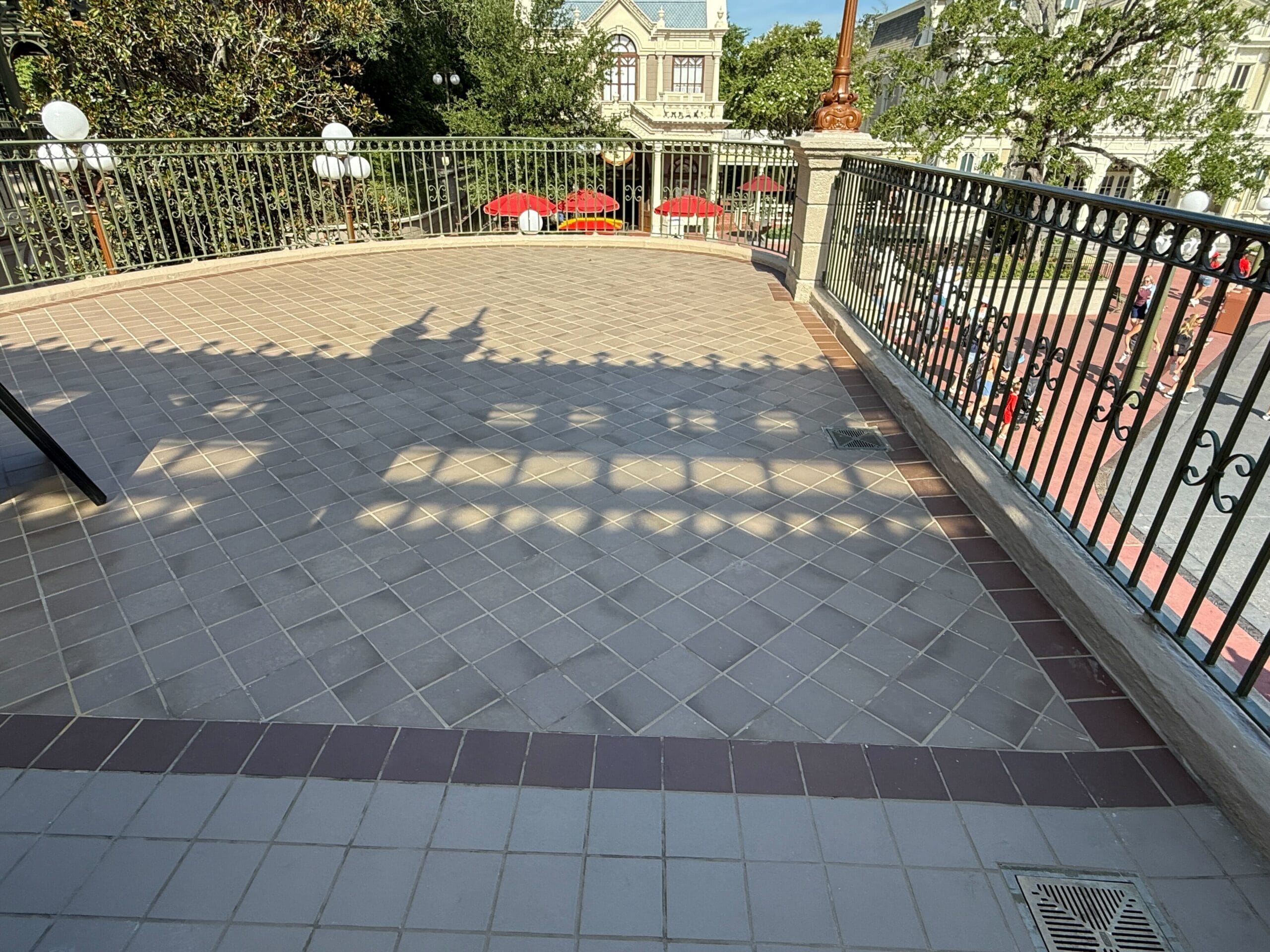 Shaded terrace with tiled floor, black railing, and red tables below in a park-like setting.
