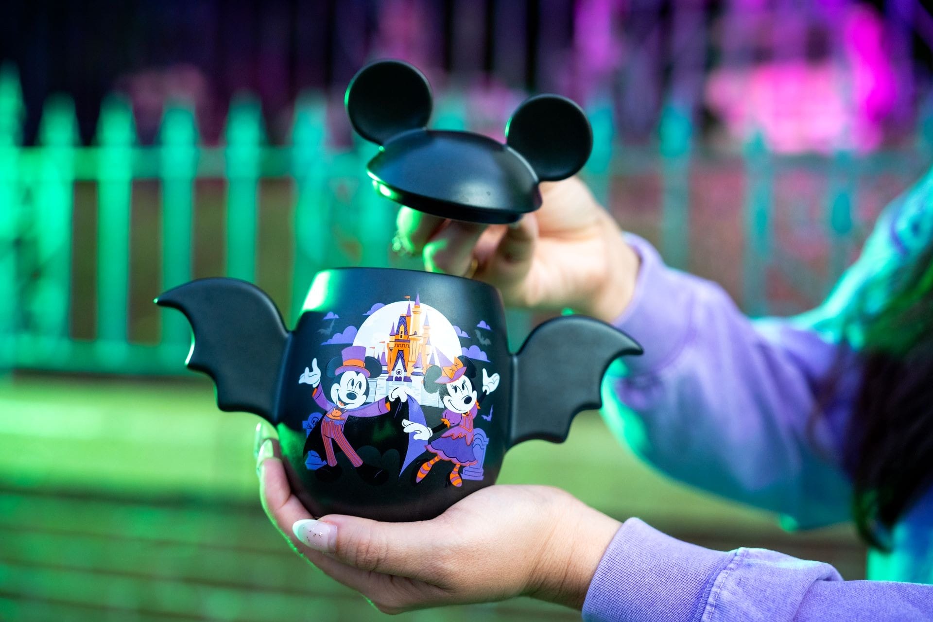 A person holds a Mickey Mouse-themed Halloween merchandise popcorn bucket with bat wings and castle art.