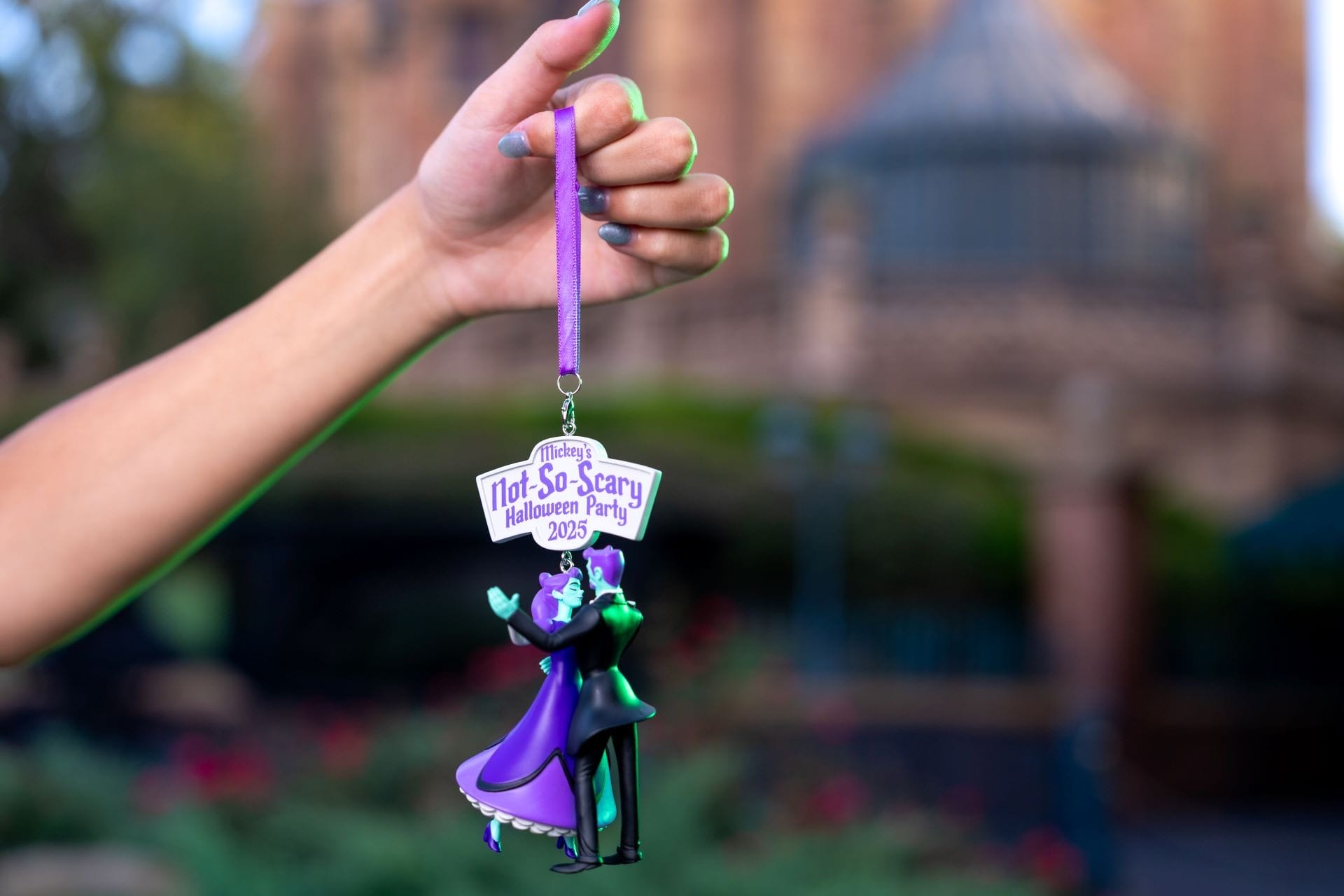 A hand holds a 2023 Mickey's Not So Scary Halloween Party ornament with two figures in purple and green outfits.