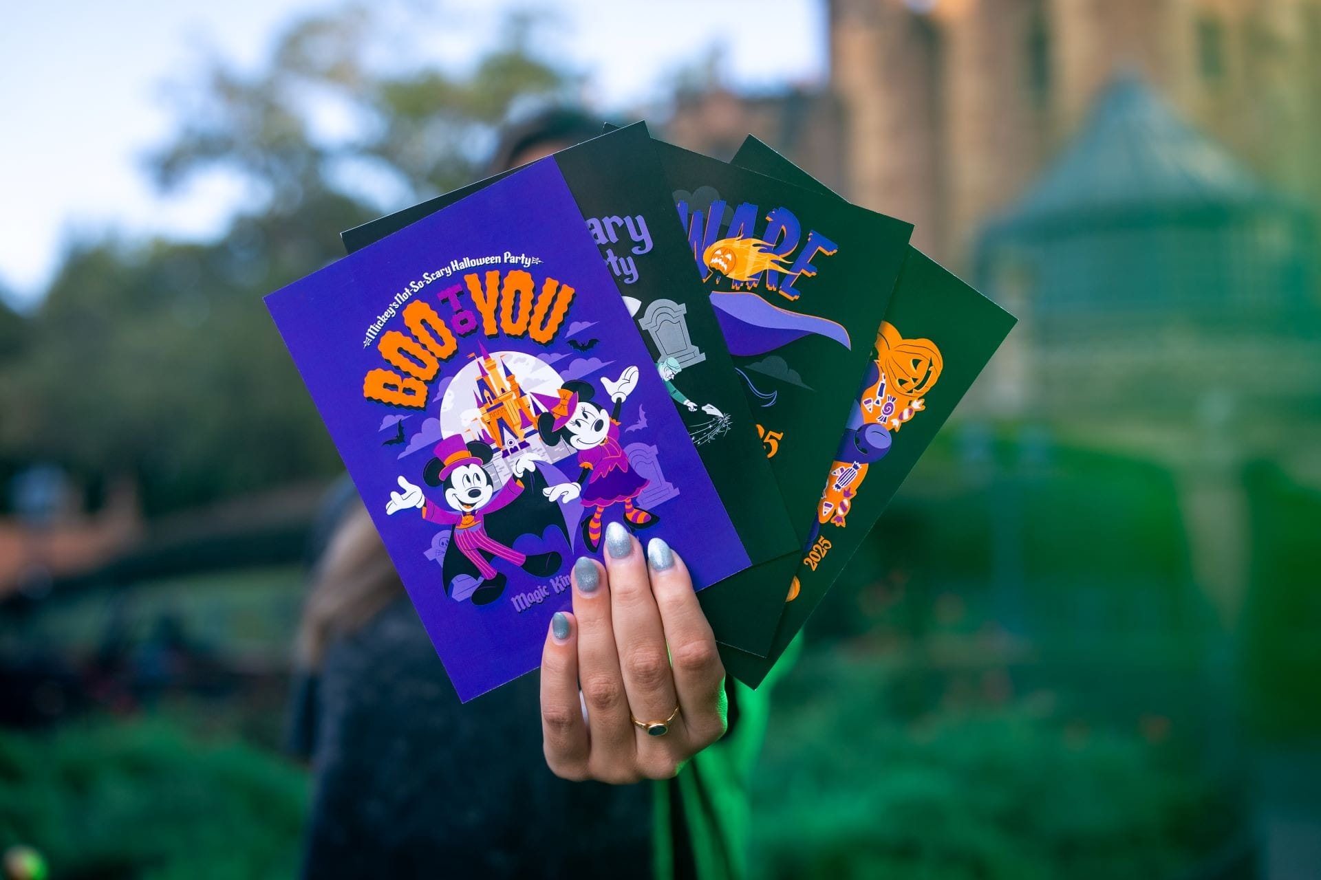 A hand with blue nails holds up four colorful Halloween-themed Disney merchandise cards featuring Mickey and friends.