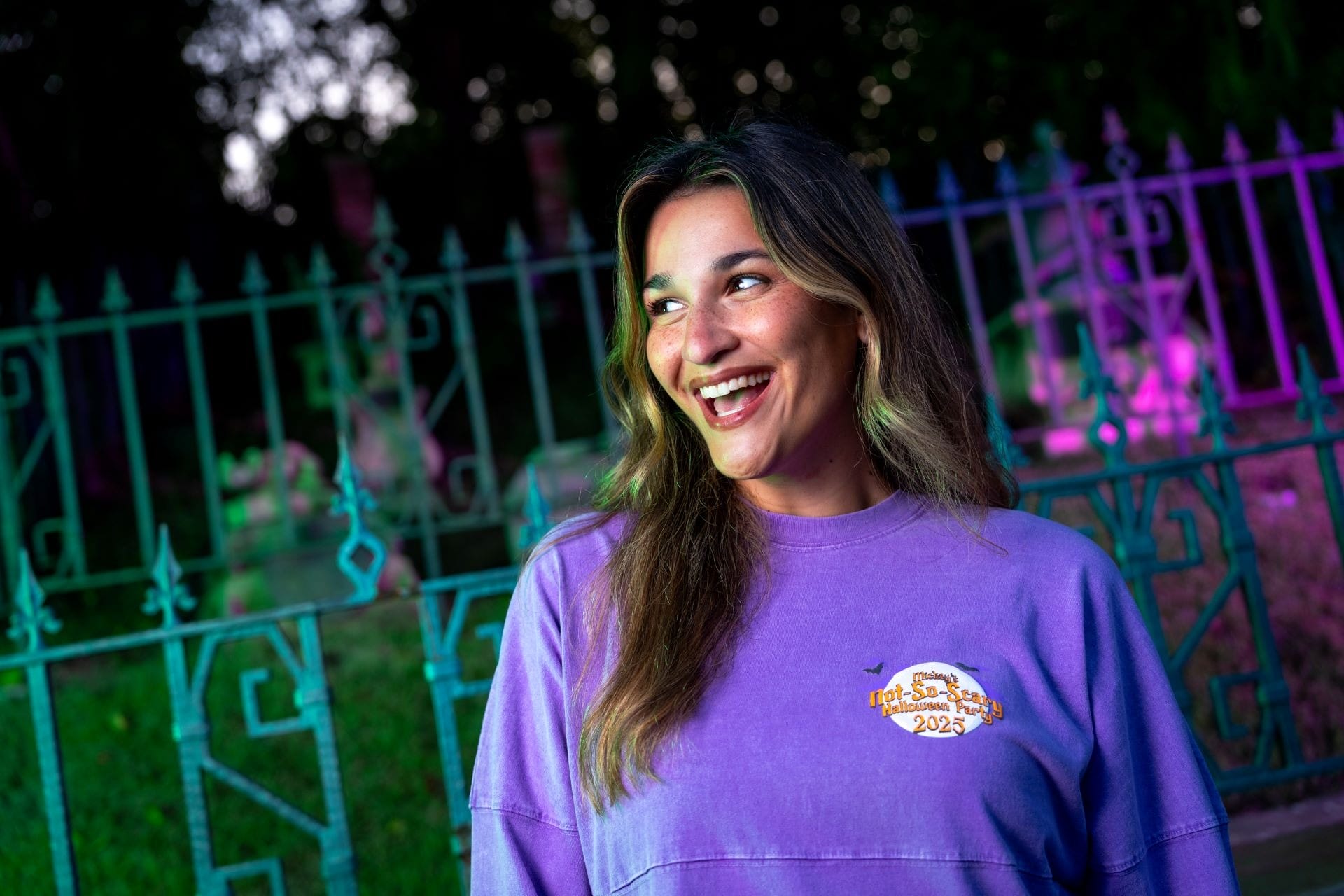 A smiling woman in a purple shirt stands by a decorative fence, illuminated by colorful Mickey's Not So Scary Halloween Party lights.