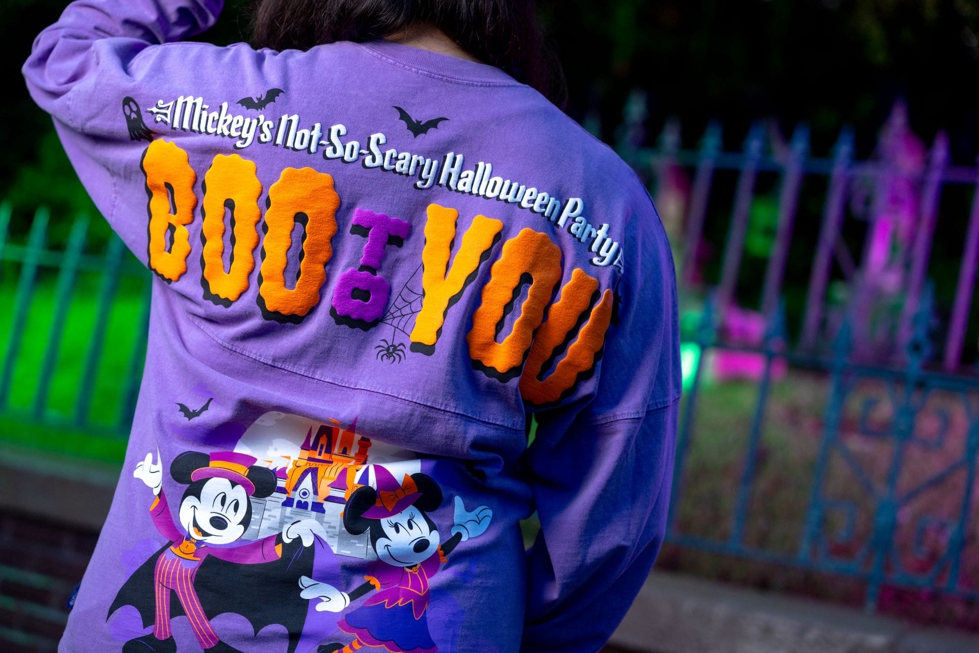 Person in a purple "Boo to You" shirt from Disney Halloween Merchandise, with Mickey and Minnie, stands by colorful party lights.