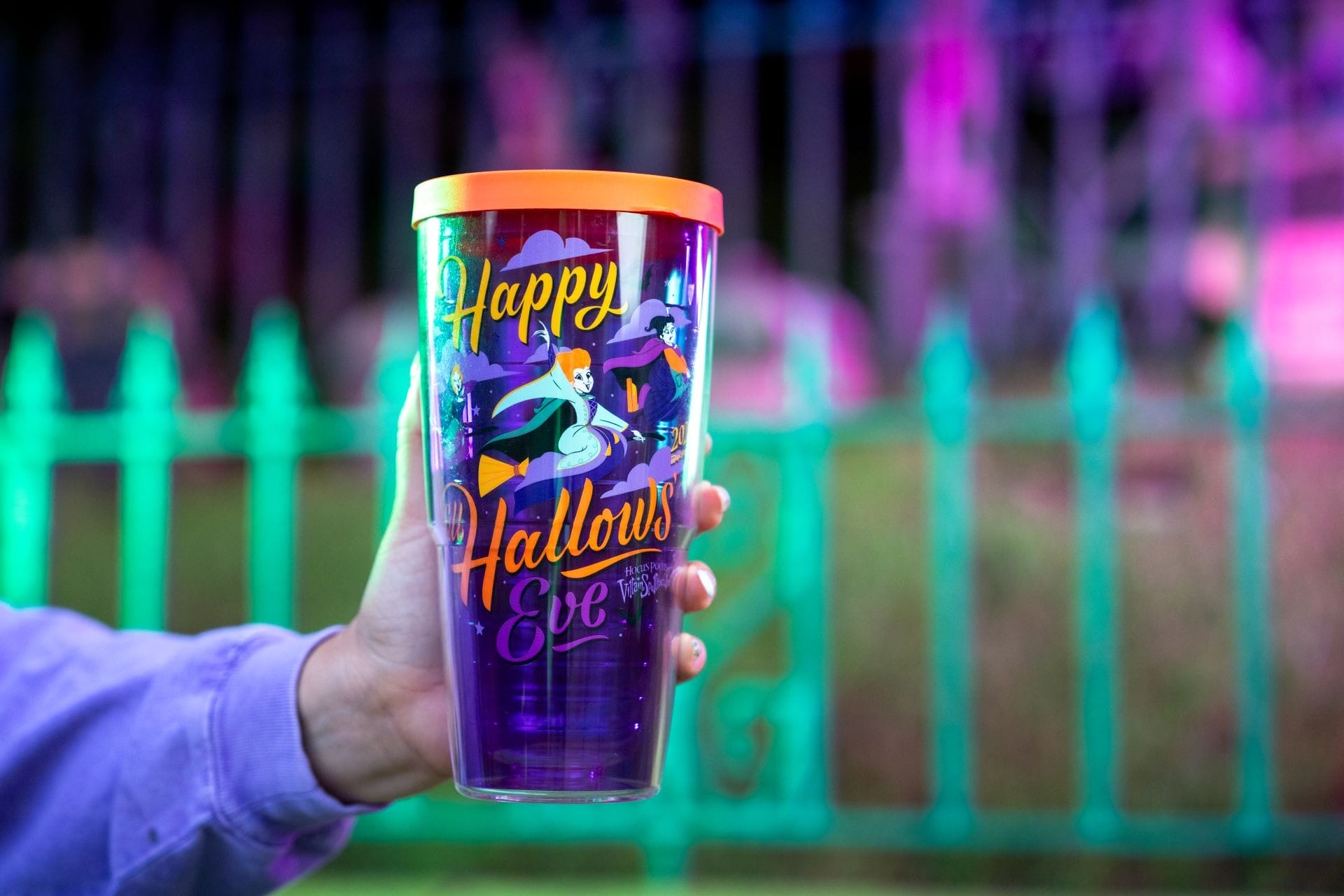 A hand holds a purple Halloween merchandise cup with cartoon ghosts and “Happy Hallows Eve” text by a colorful fence.