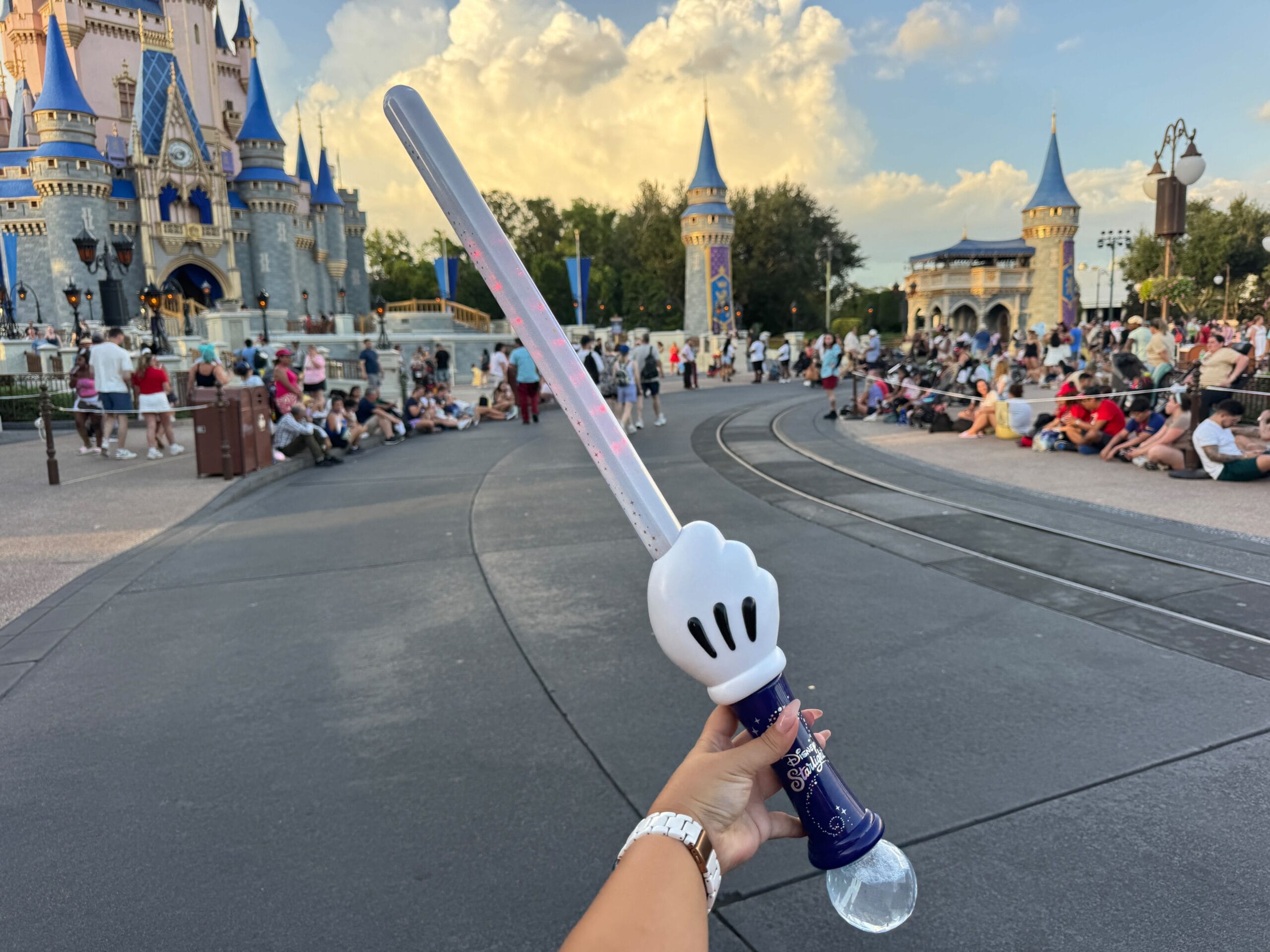 A hand holds a Mickey Mouse glove-shaped light-up wand in front of a Disney castle with crowds nearby.