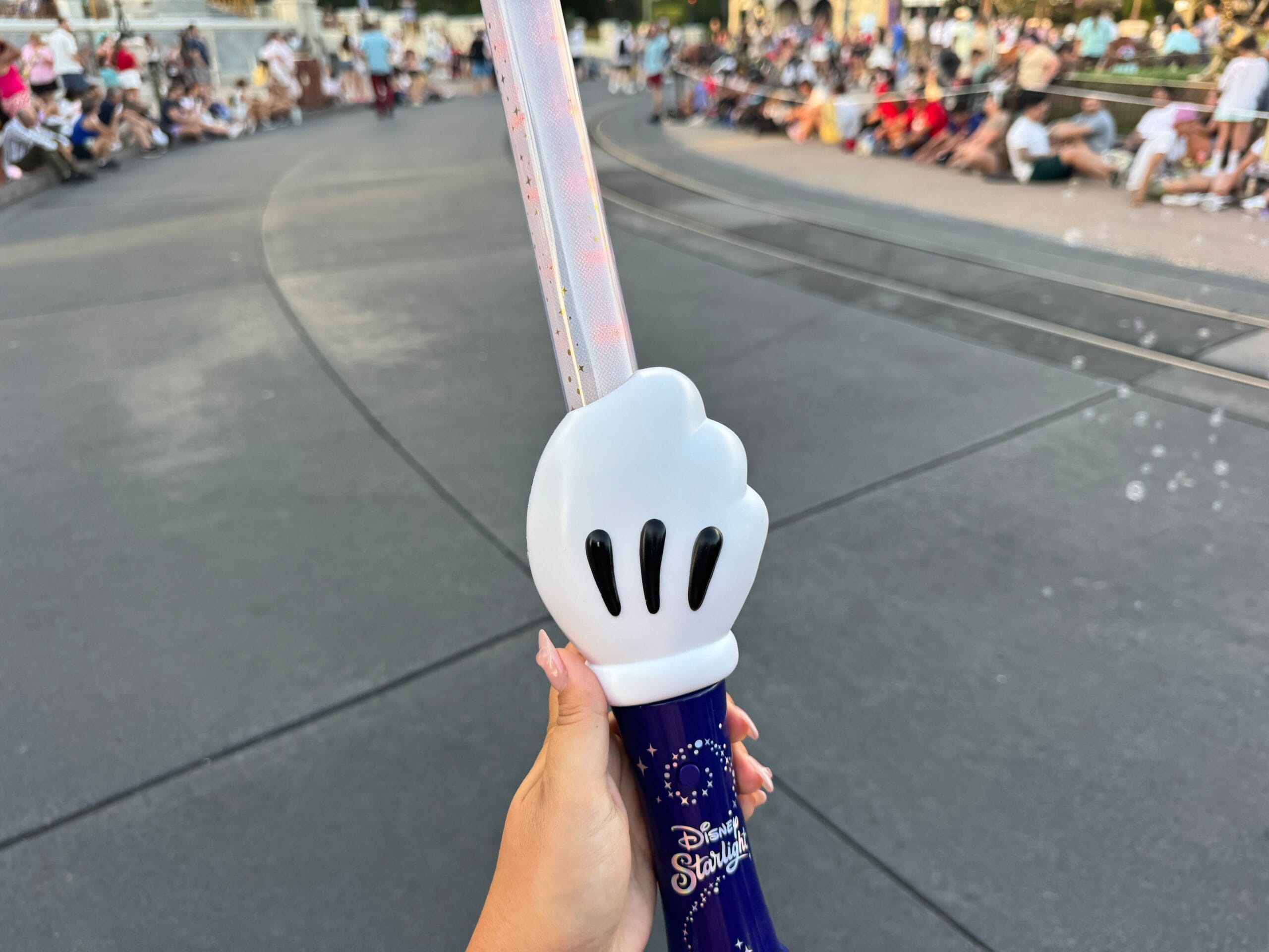 A hand holds a Disney light-up wand shaped like a white glove, with people sitting along a parade route in the background.