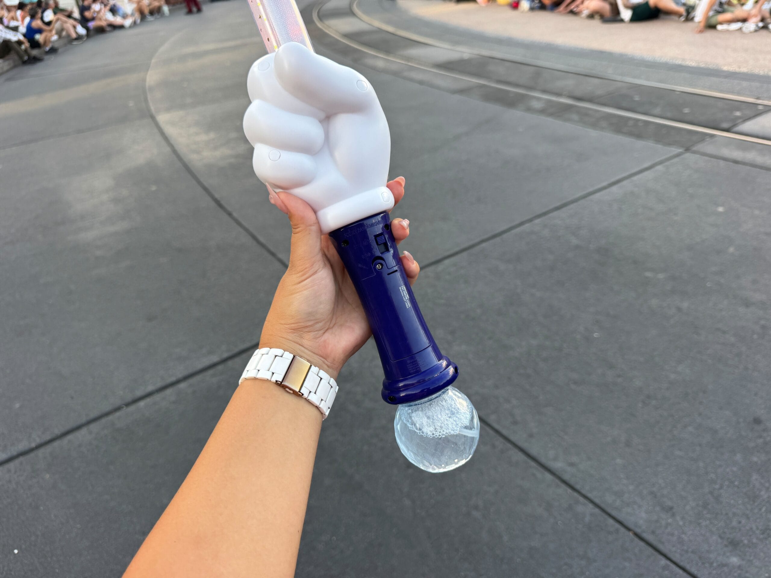 A hand holds a Mickey Mouse glove-shaped light wand with people sitting along a parade route in the background.