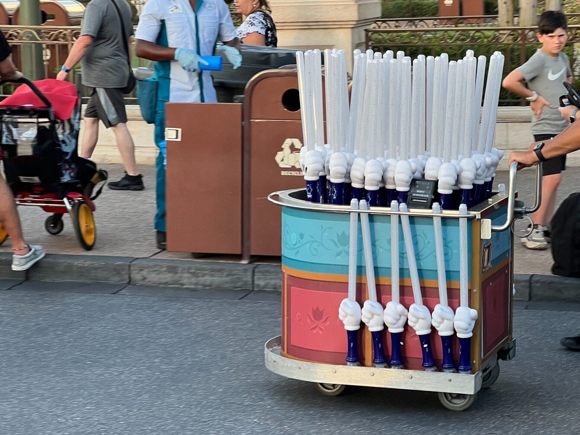 A cart full of toy lightsabers is pushed along a theme park path with people and a trash bin nearby.