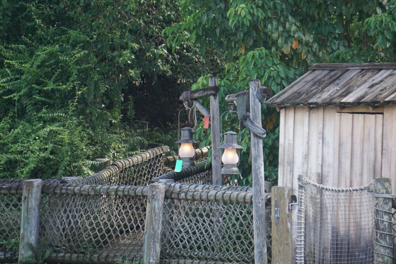 Two lanterns hang by a rope bridge near a wooden shed, surrounded by dense green foliage.