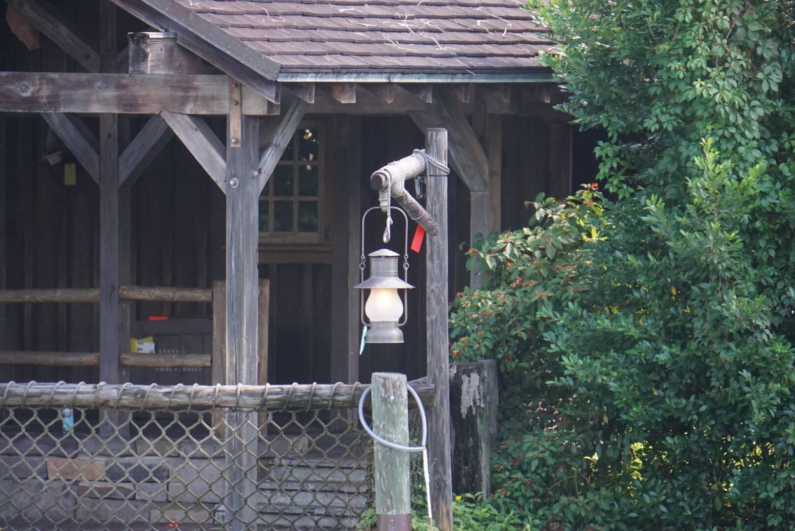 A lantern hangs from a rustic post in front of a wooden cabin surrounded by greenery.