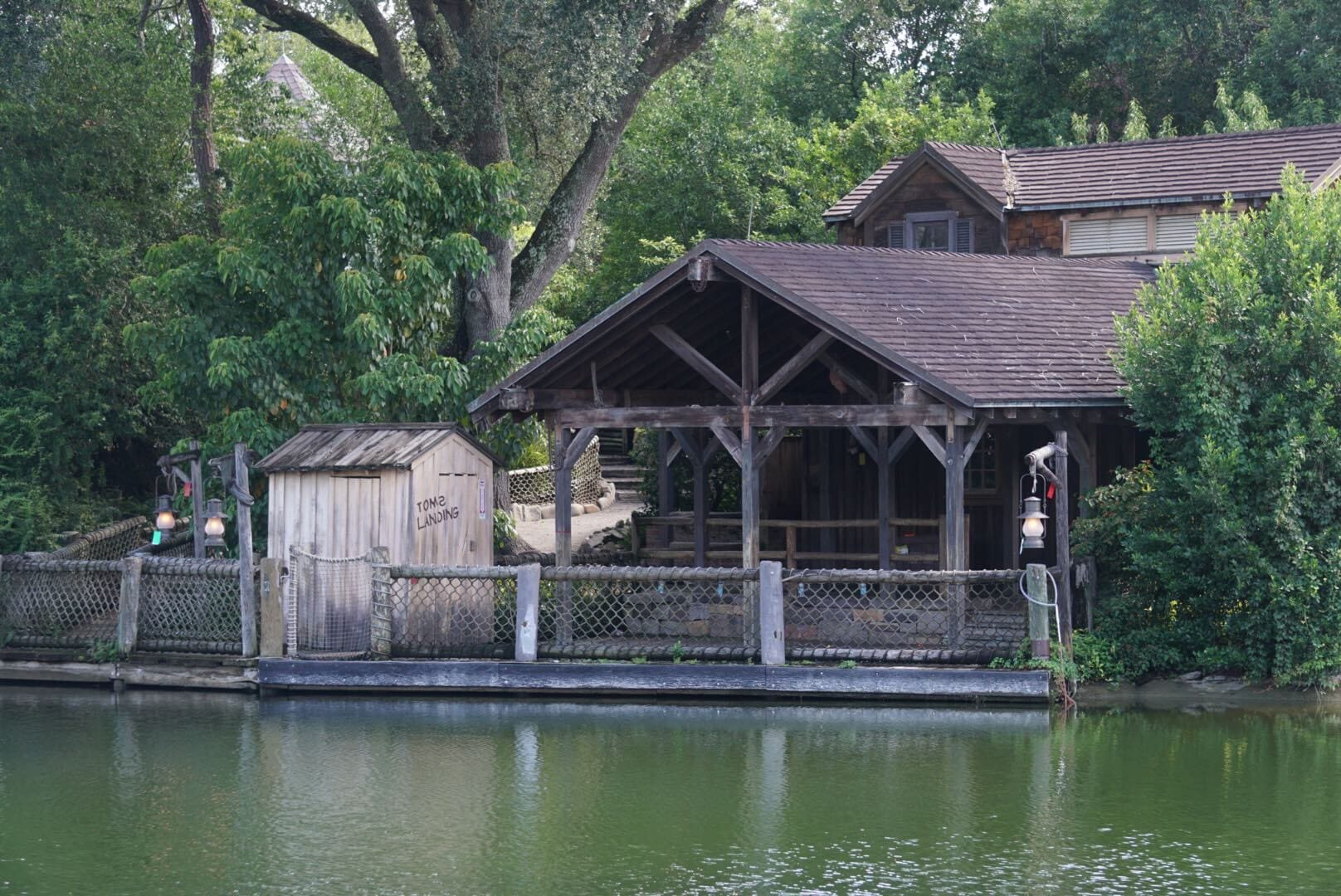 A rustic wooden boathouse and shed sit by a calm river, surrounded by lush green trees and foliage.
