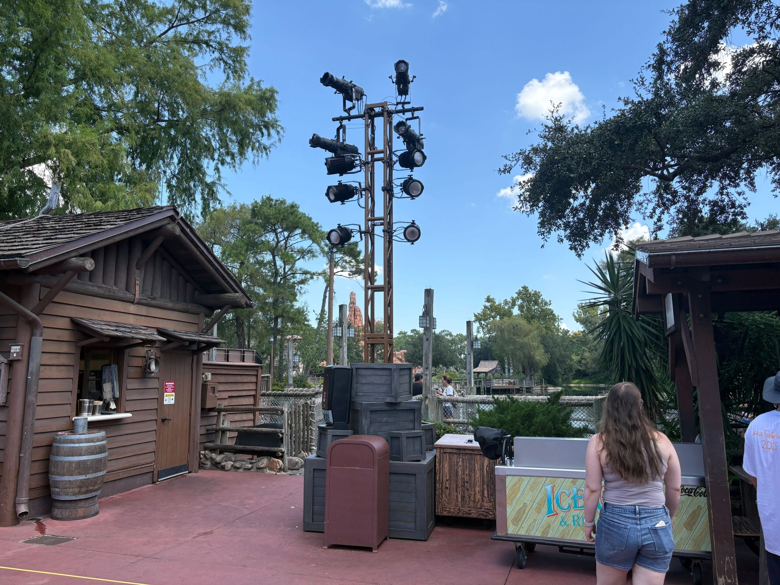 A woman stands near an ice cart by the lakeside, with Magic Kingdom’s new trusses and rustic wooden buildings behind.