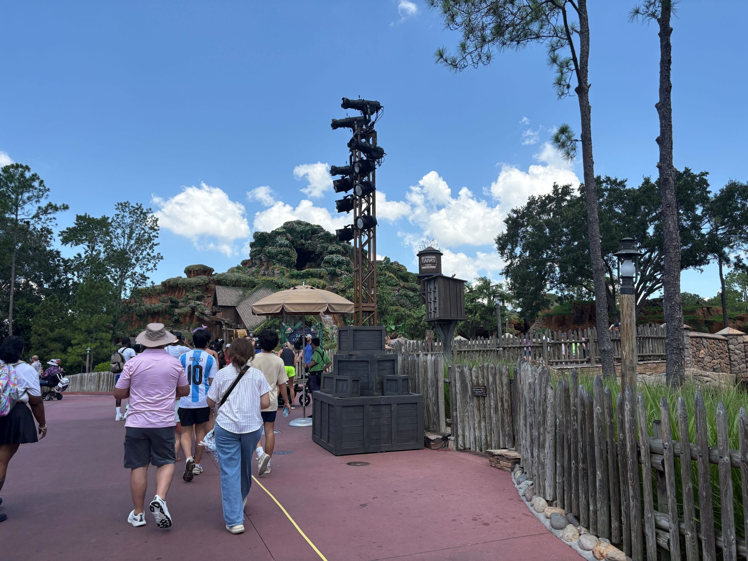 People stroll near a wooden fence at Magic Kingdom, with trees and rocky structures under a clear blue sky.