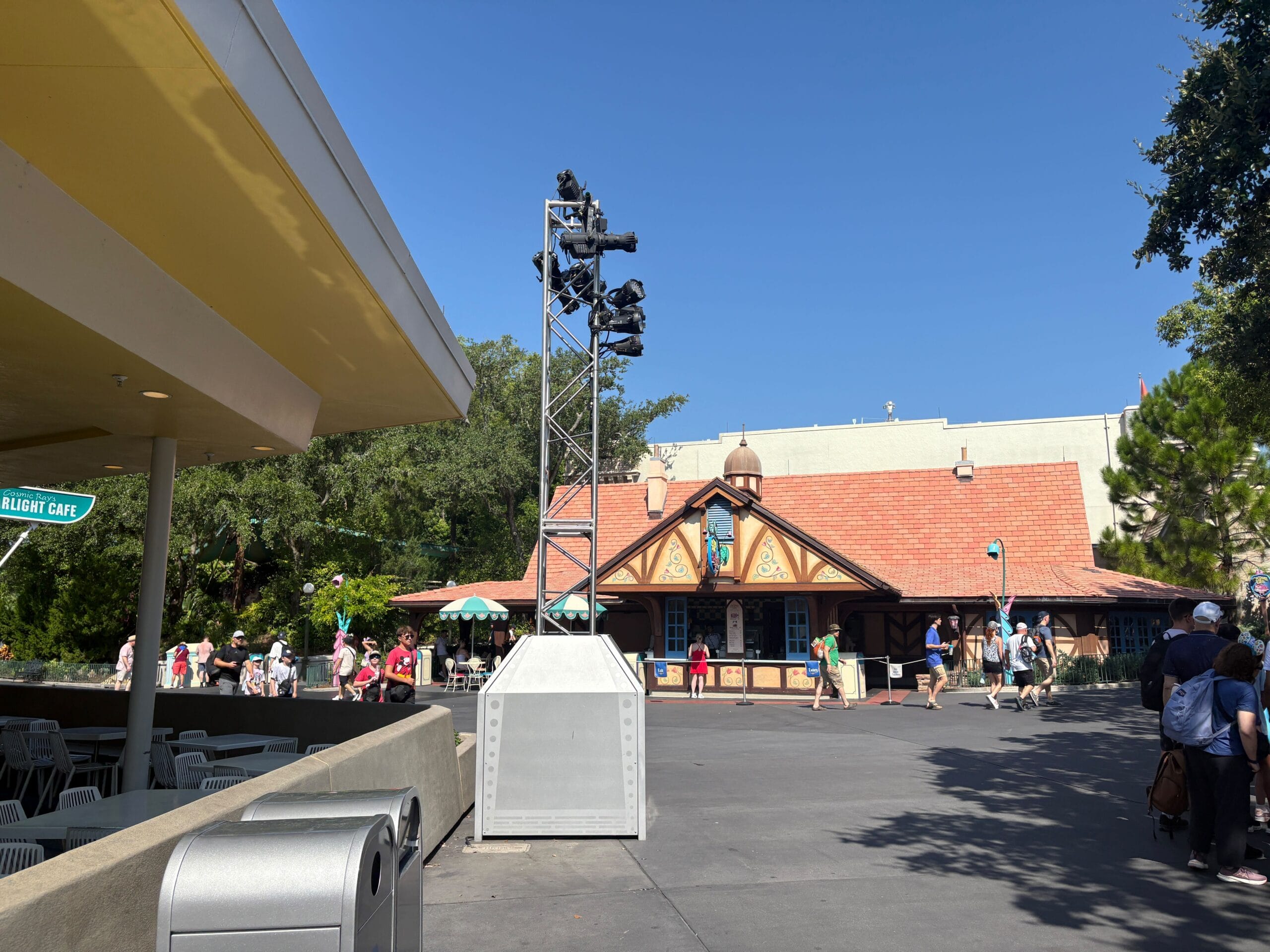 People walk near Cheshire Cafe at Magic Kingdom, with trusses and lighting visible in the background.