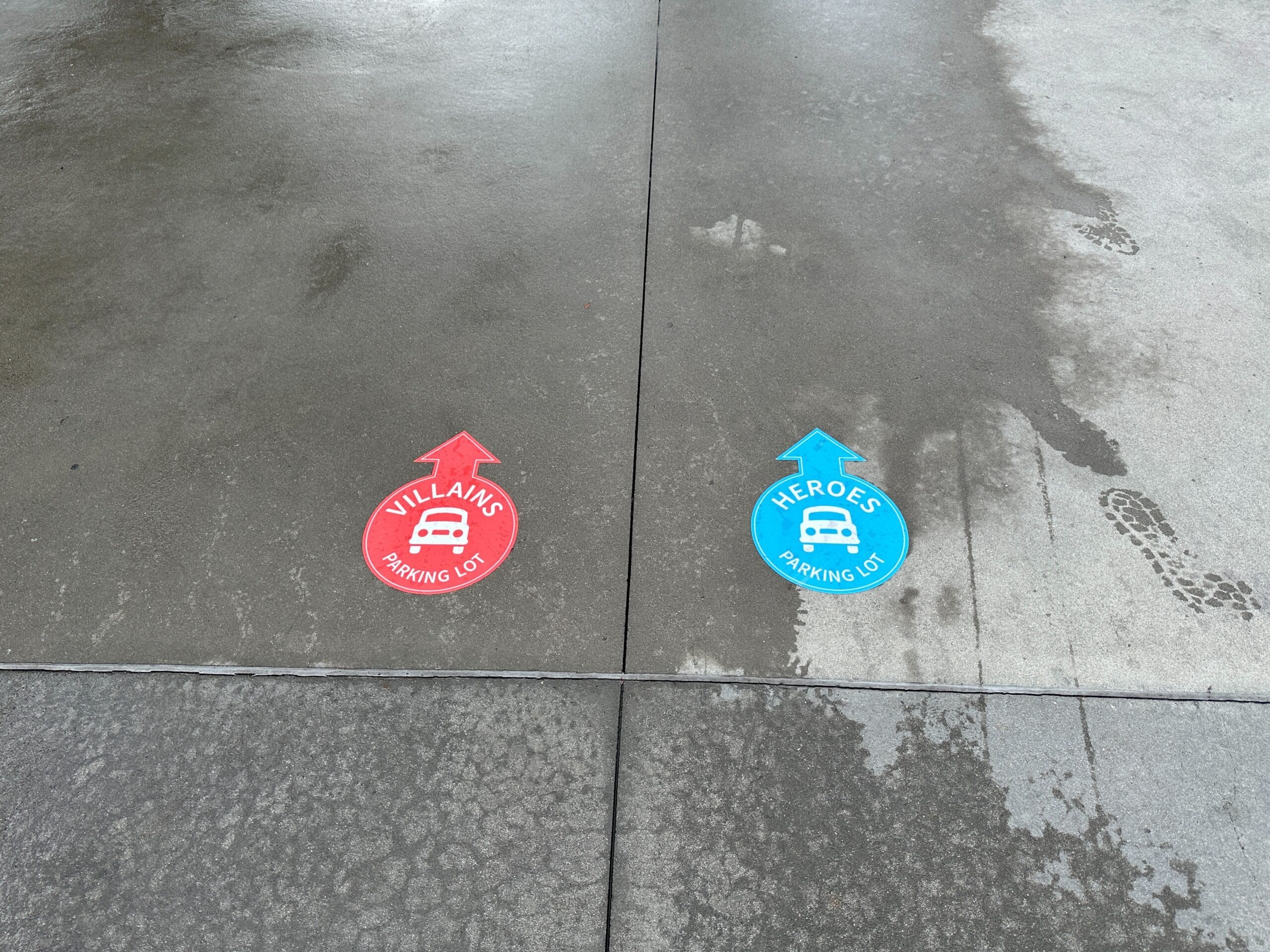 Red "Villains" and blue "Heroes" parking lot signs with arrows, labeled on wet concrete pavement.