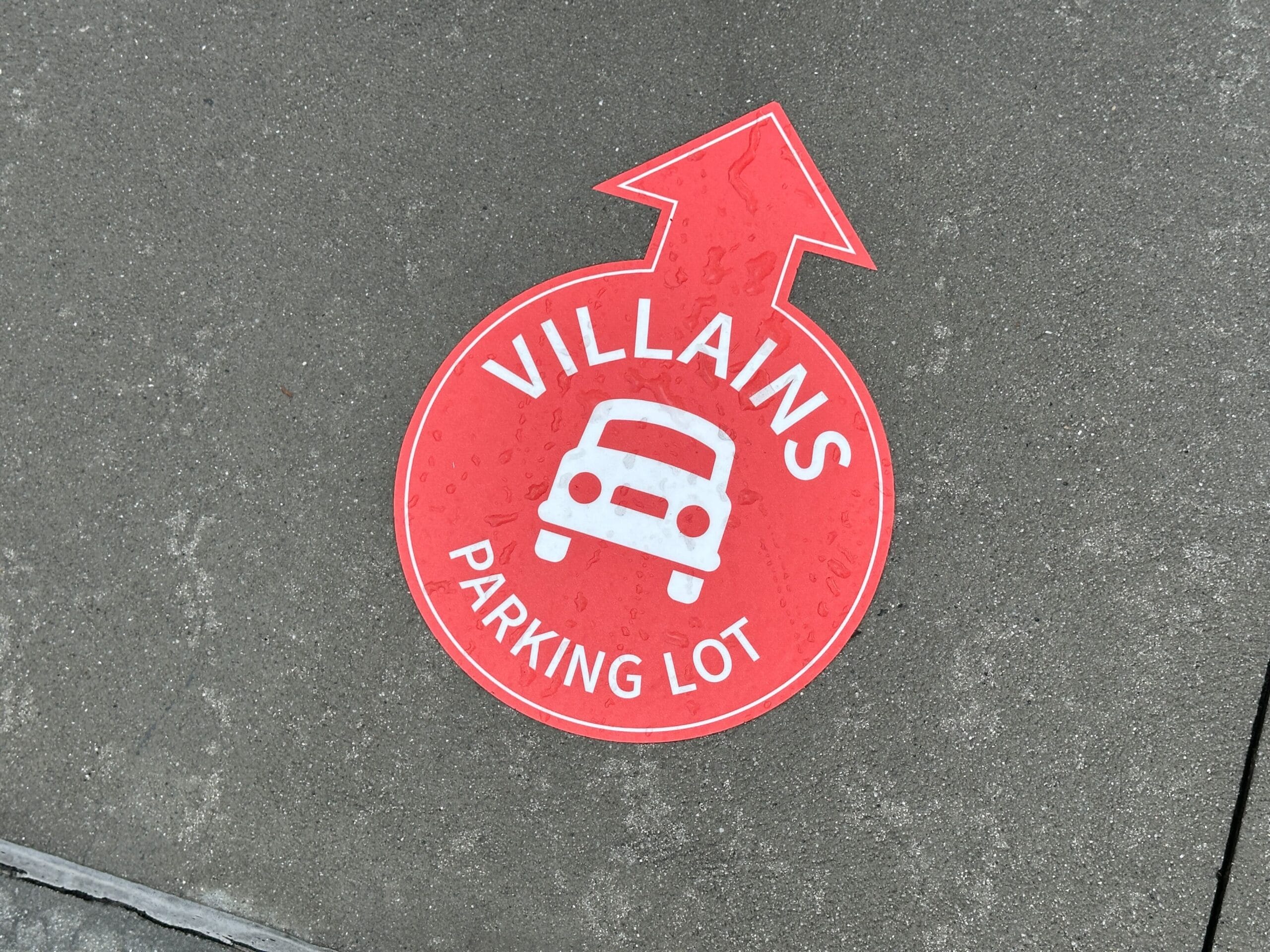 Red floor sign with an arrow, a car icon, and the words "VILLAINS PARKING LOT" pointing forward.