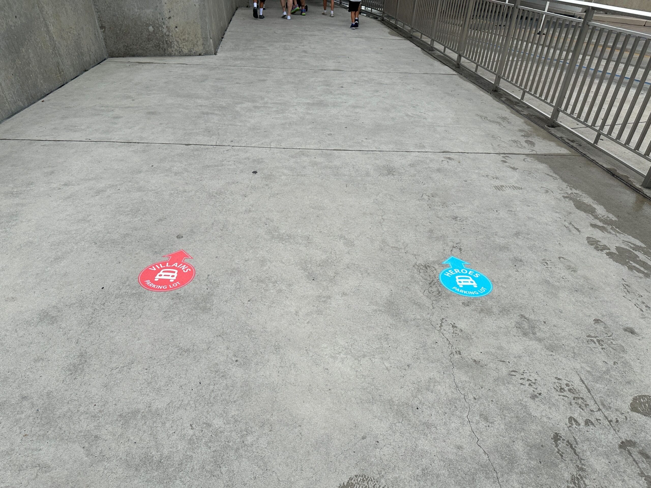 Two round floor signs mark "Villains" in red and "Heroes" in blue on a concrete walkway near metal railings.