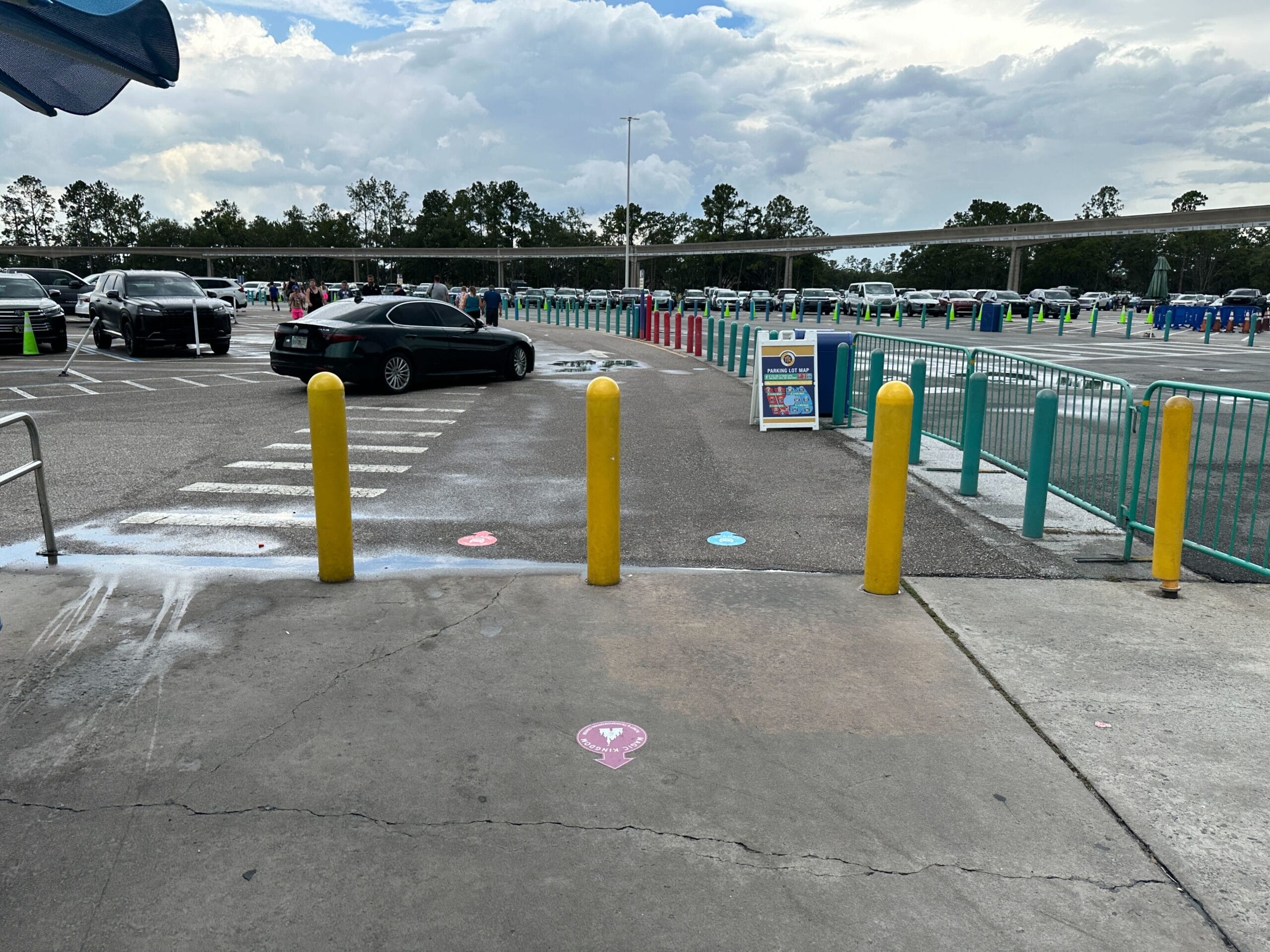Parking lot with yellow bollards, crosswalk, Parking Lot Signs; cars and trees in the background under a cloudy sky.