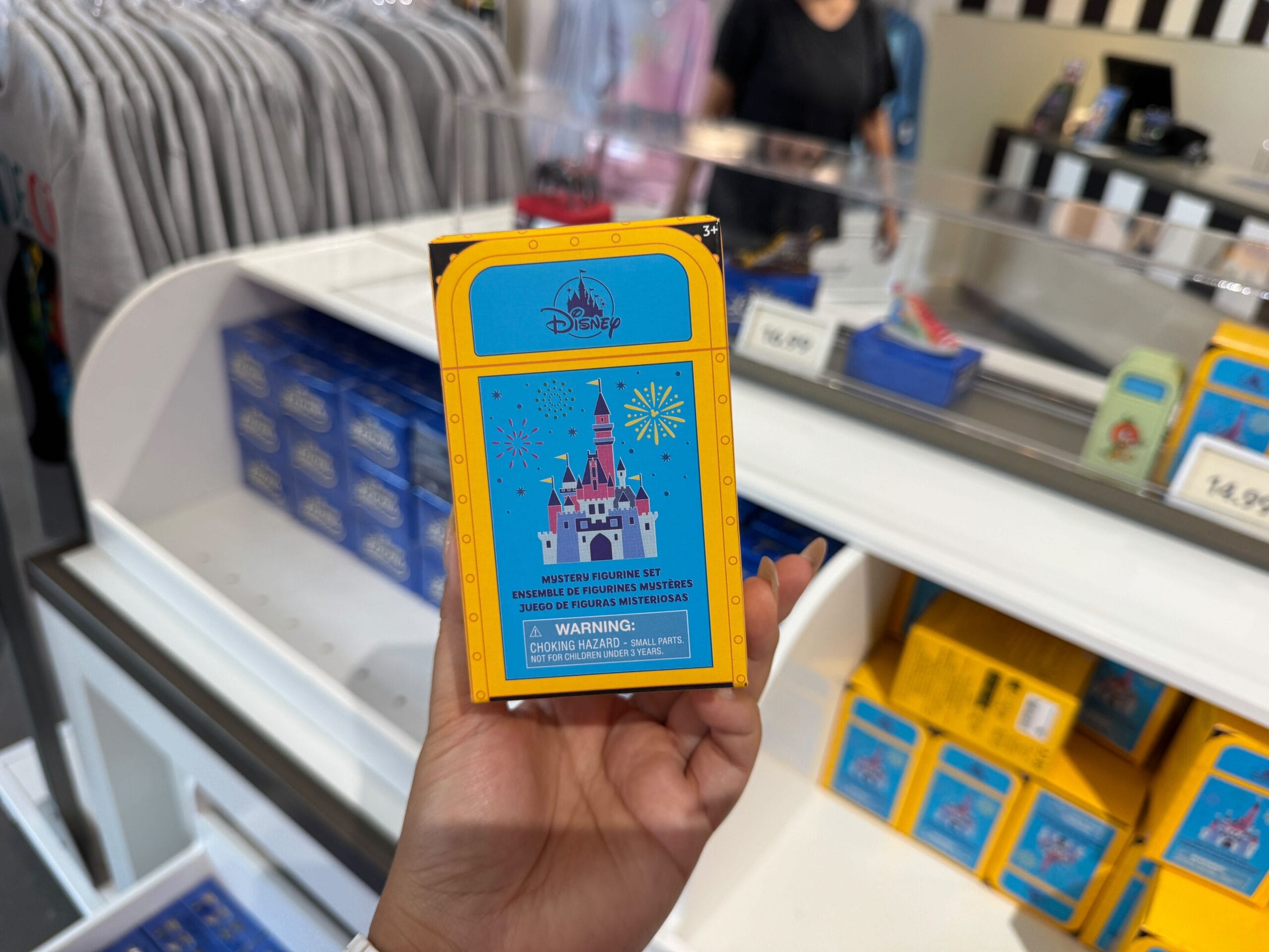 Hand holding a Disney-themed mystery pin box featuring a pink castle, fireworks, and a warning label in a store.
