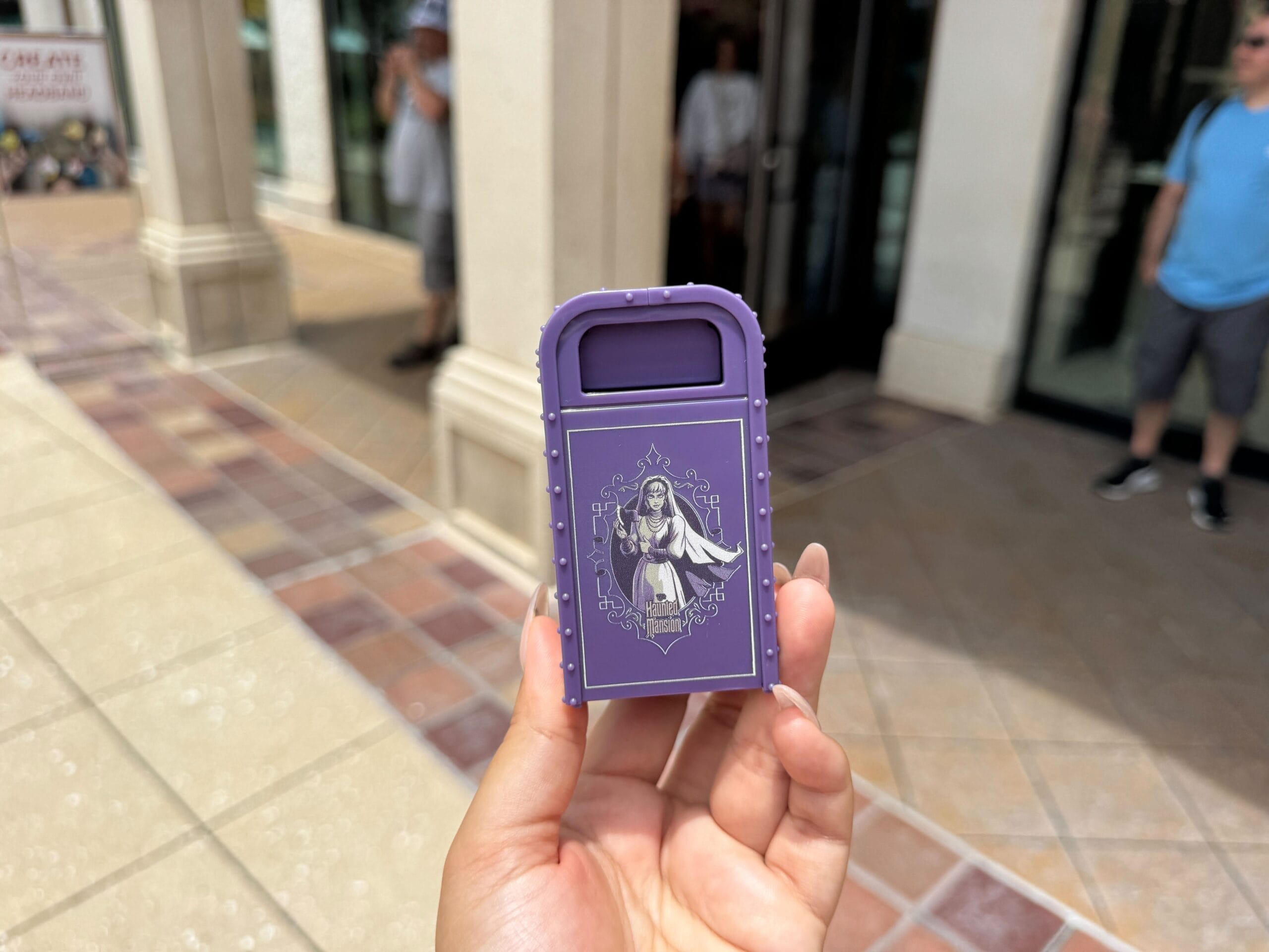 A hand holds a small purple trash can with a spooky illustration, outdoors near a building entrance.