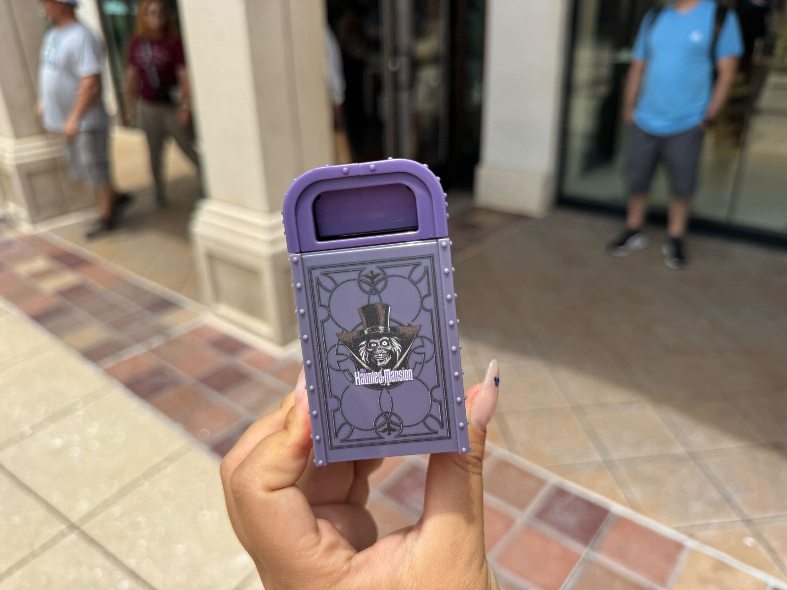 A hand holds a small purple Haunted Mansion-themed trash can souvenir outside a building.