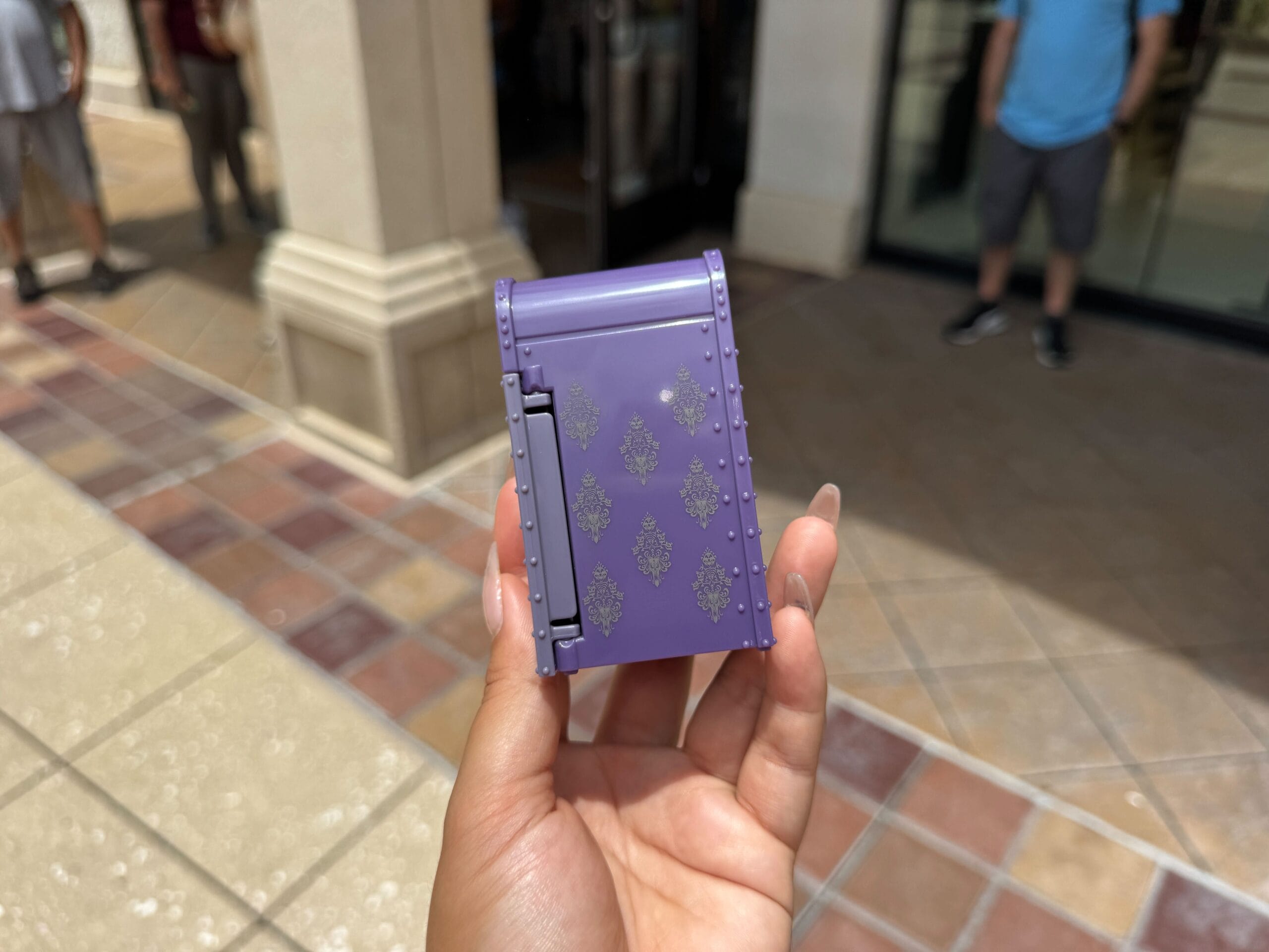 A hand holds a small purple, patterned box outdoors near a building entrance.