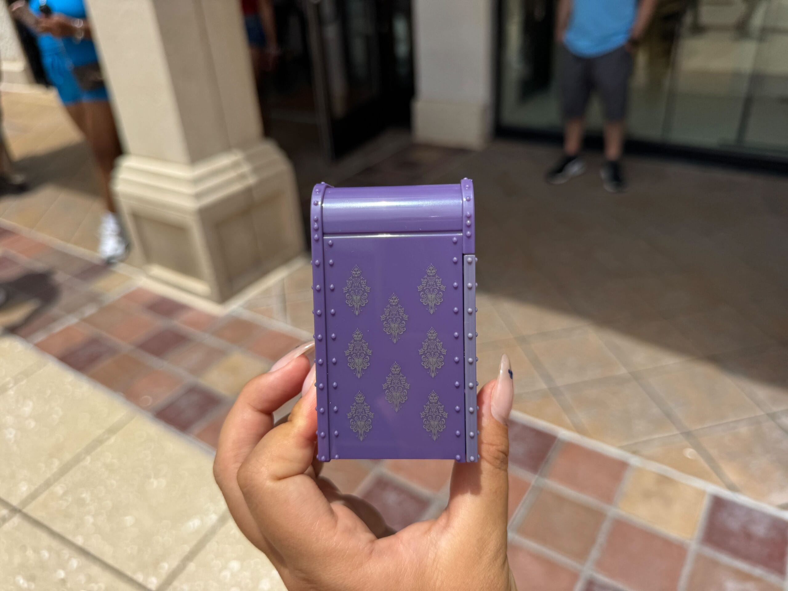 A hand holds a small, purple, decorative box outdoors on a tiled pavement.