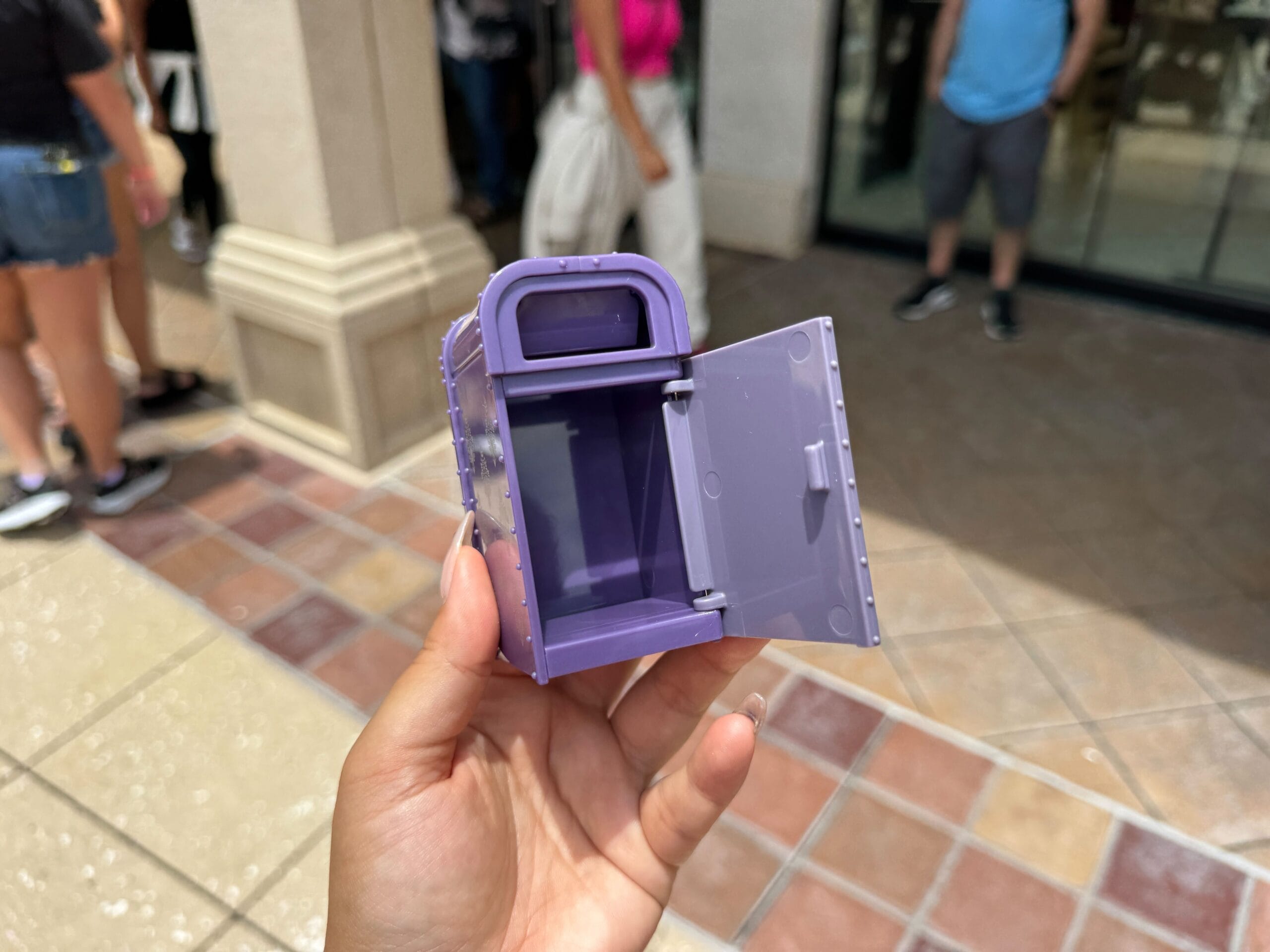 A hand holds a small, open, purple mailbox toy outdoors with people standing in the background.