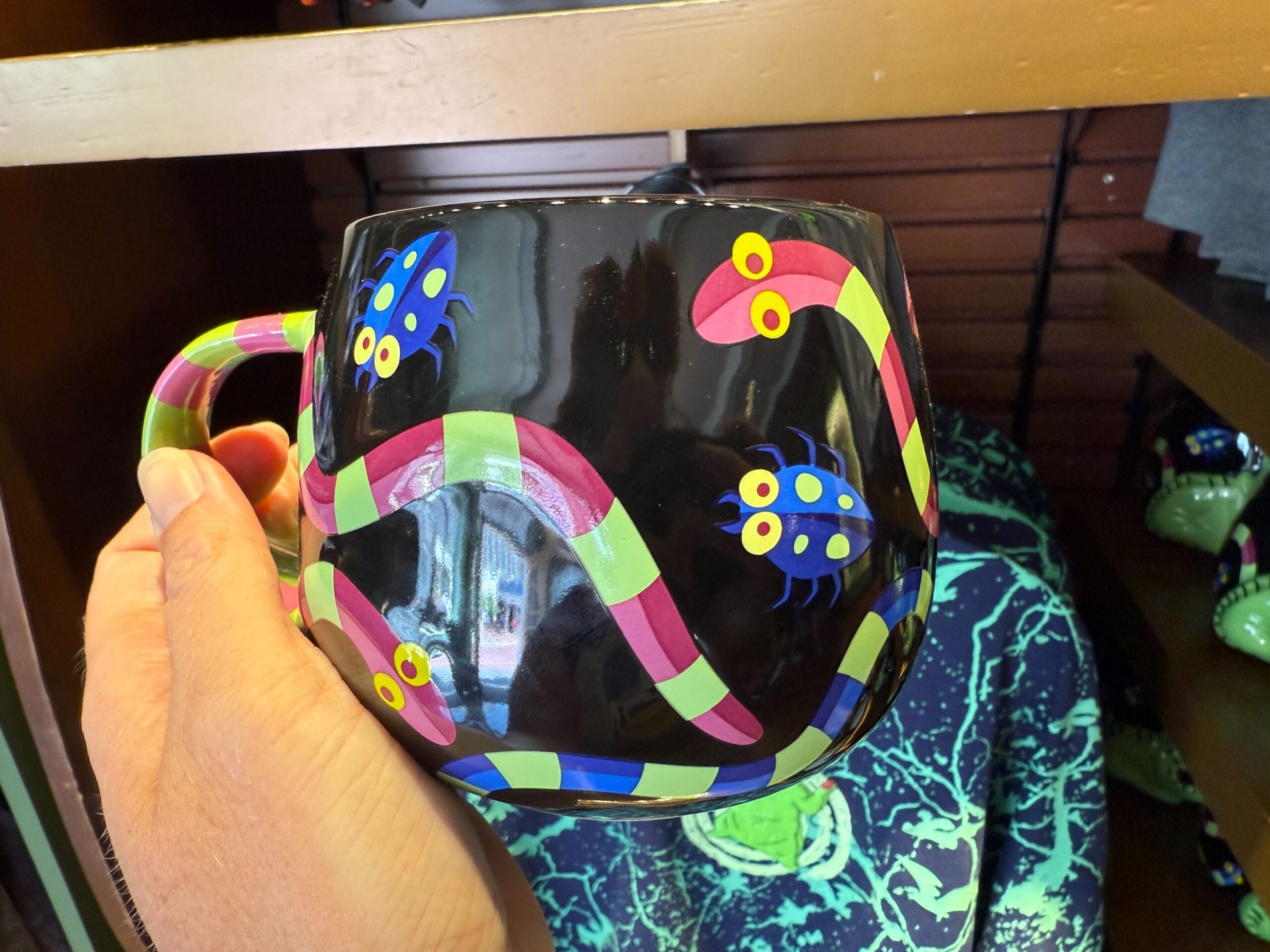 A hand holds a black mug with colorful cartoon snakes and bugs—new merchandise from The Nightmare Before Christmas.