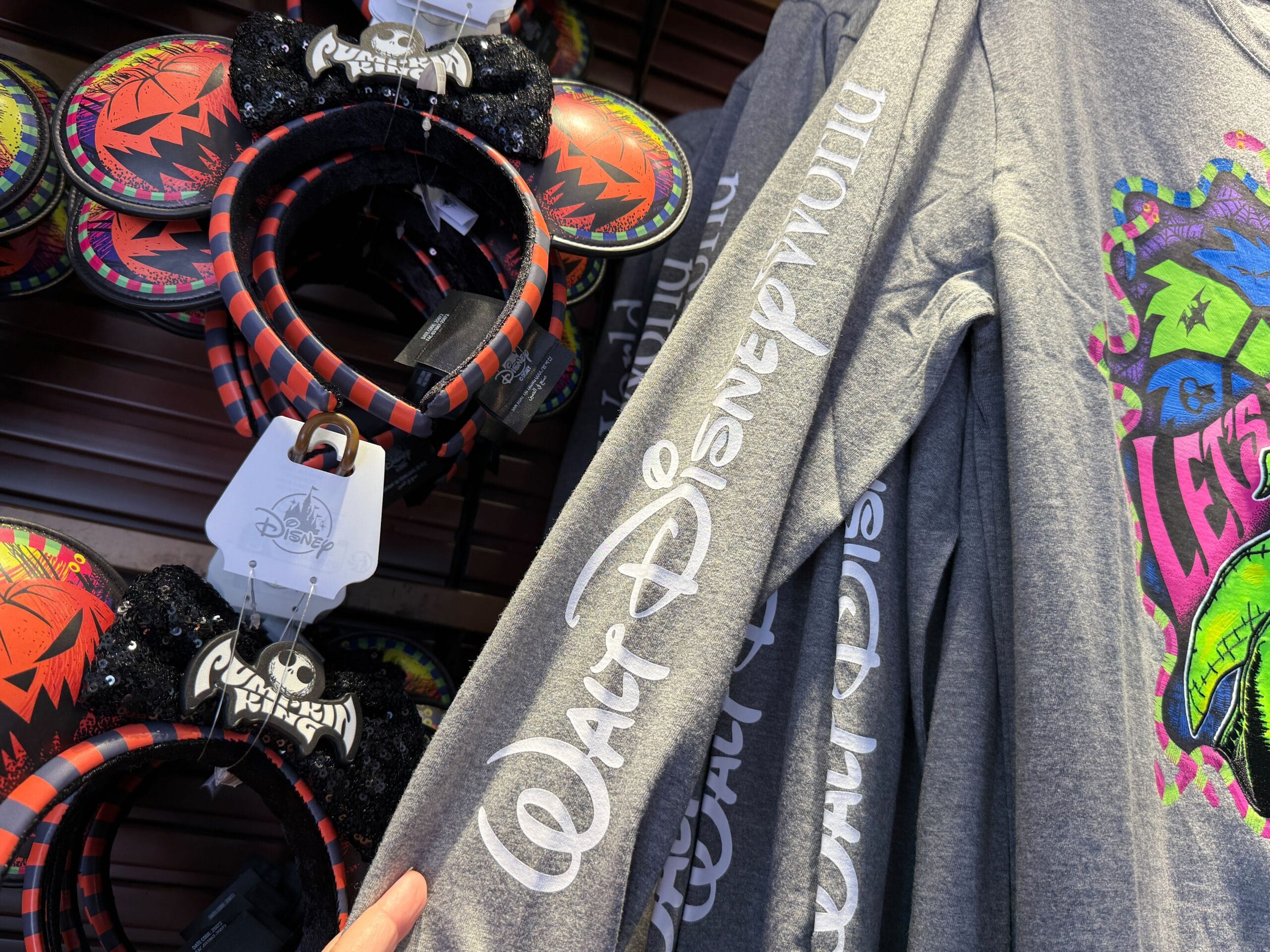 A hand holds a gray Walt Disney World shirt near Nightmare Before Christmas merchandise and villain Mickey ear headbands.