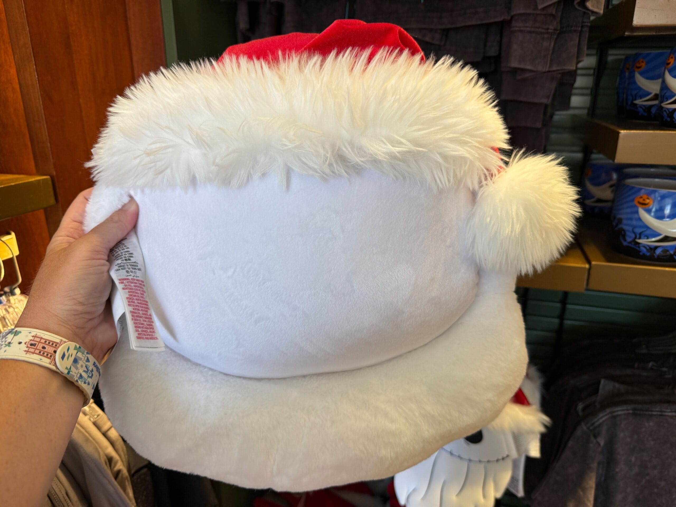 A hand holds a plush white Santa hat with fluffy trim, featured among Nightmare Before Christmas merchandise in a store display.
