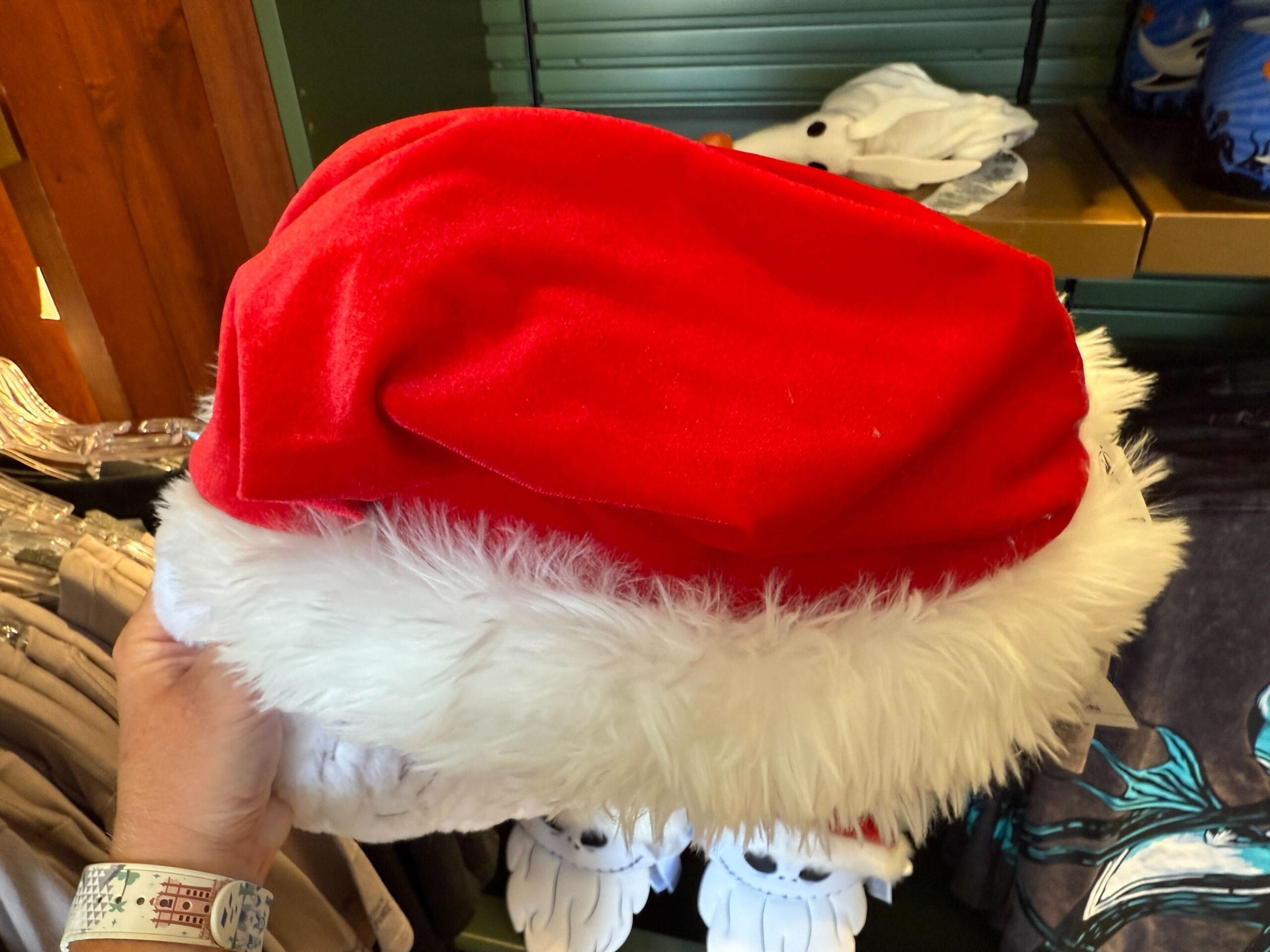 A hand holds a red Santa hat with white fur trim, browsing holiday merchandise on store shelves in the background.