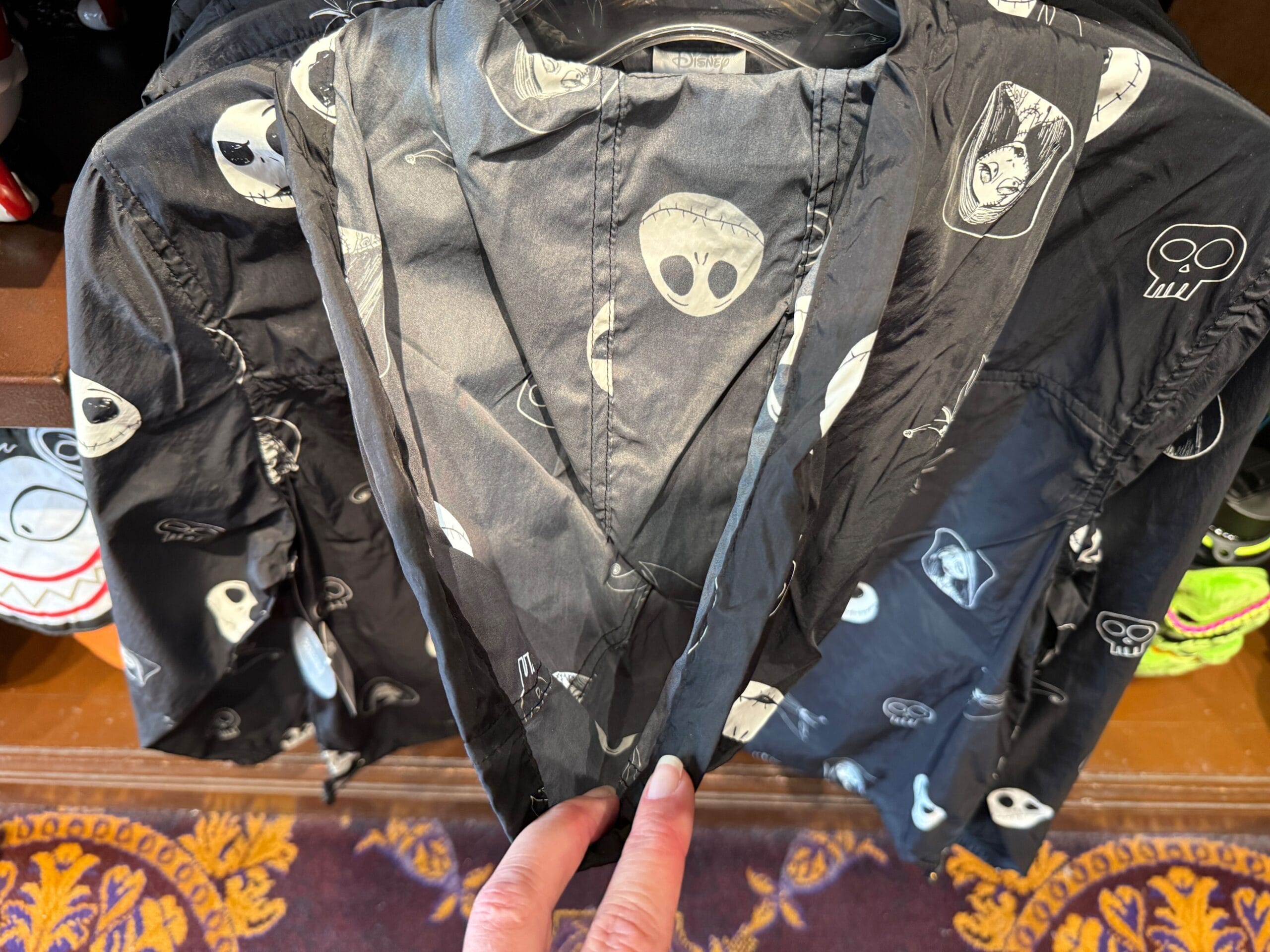 A hand holds new Nightmare Before Christmas merchandise: a black jacket with white alien faces and skulls over a carpet.