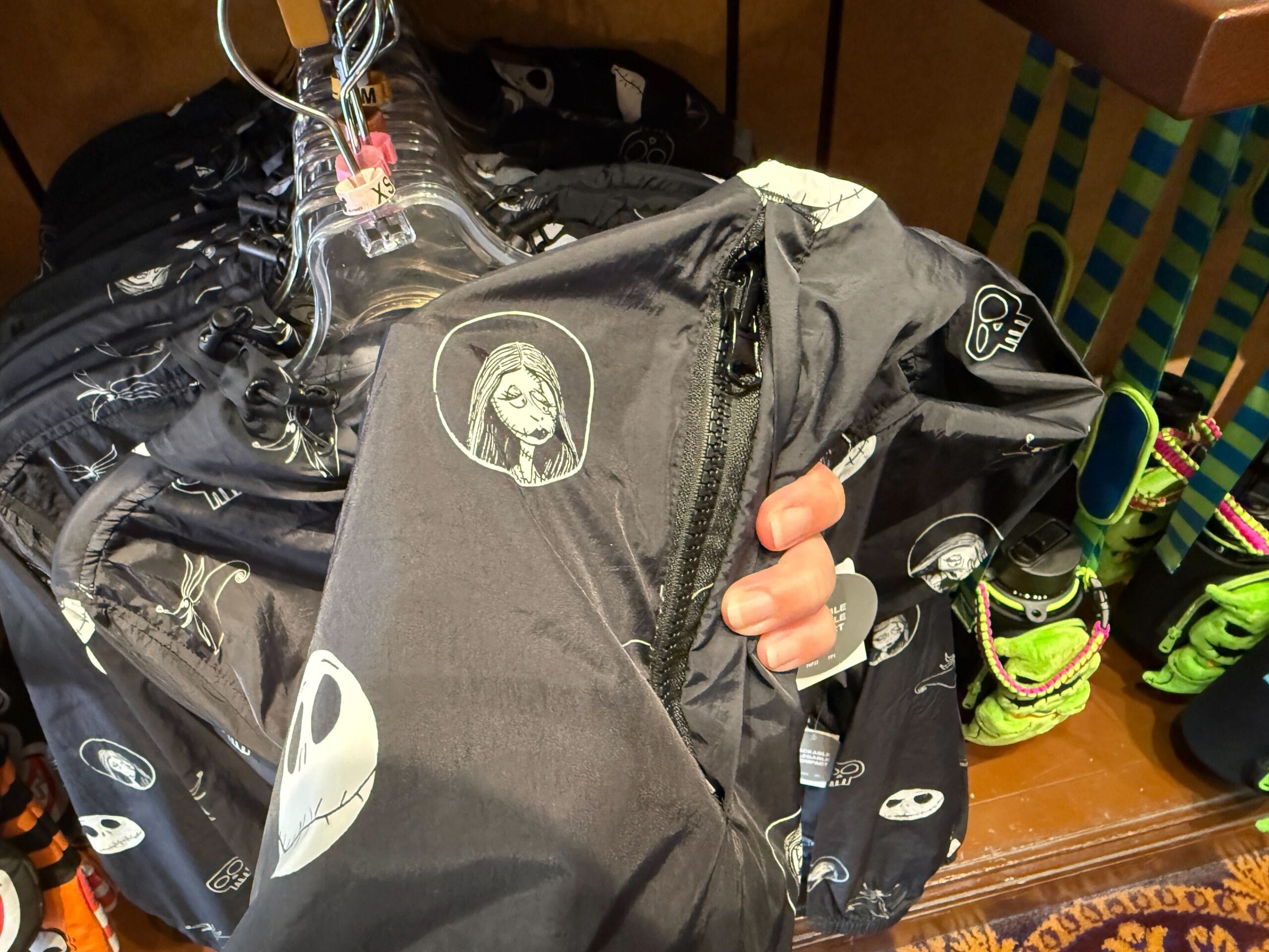 A hand holds a black new Nightmare Before Christmas jacket with white characters, hanging on a rack.